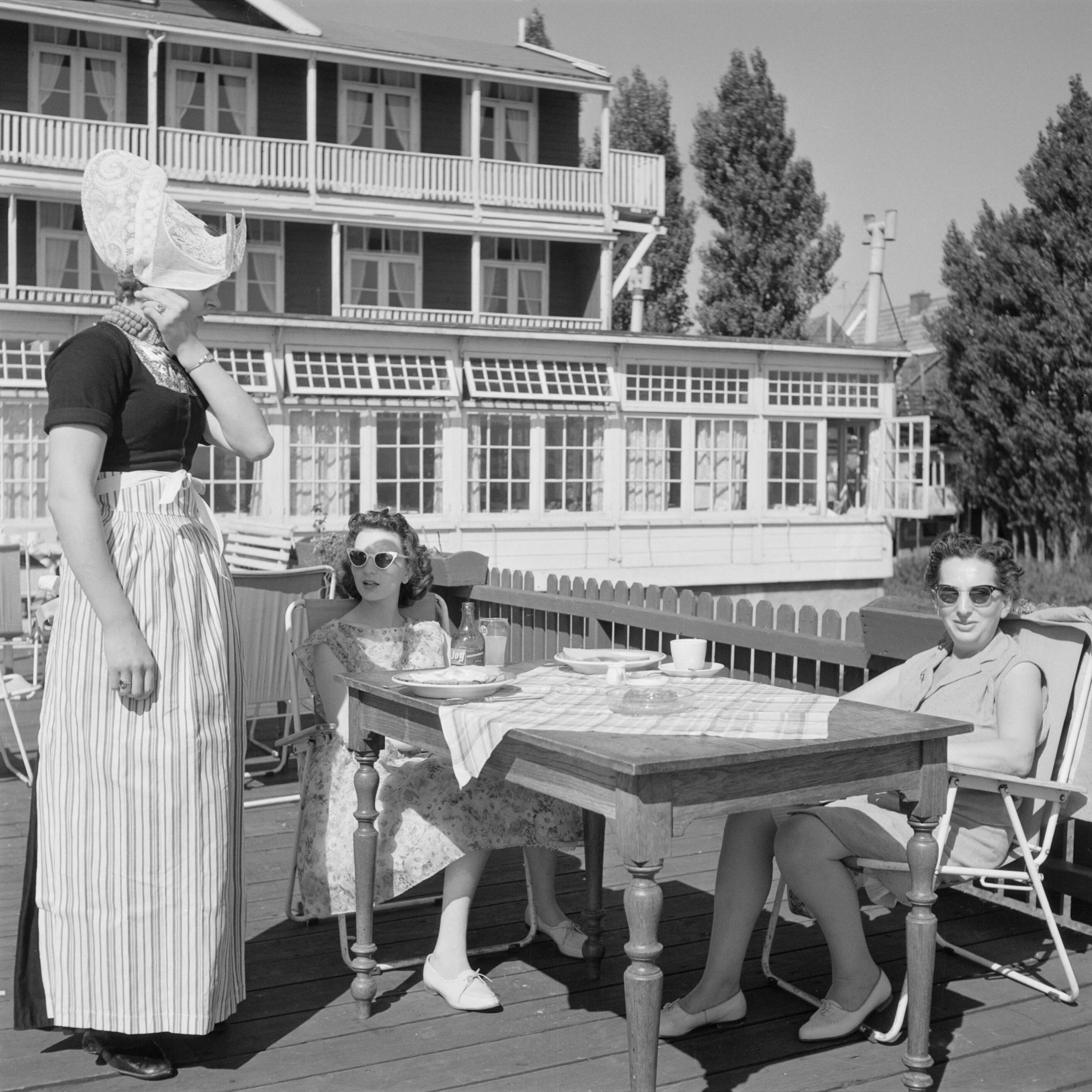 Volendam — Volendam. Een medewerkster van Hotel Spaander bedient in klederdracht klanten (rechts: Hildegard van de Poll-Eschen, de echtgenote van de fotograaf) op een terras — Foto: Poll, Willem van de. Licentie: Set B: Rechtenvrij / Publiek Domein. Bron: Nationaal Archief. http://hdl.handle.net/10648/aecedc84-d0b4-102d-bcf8-003048976d84