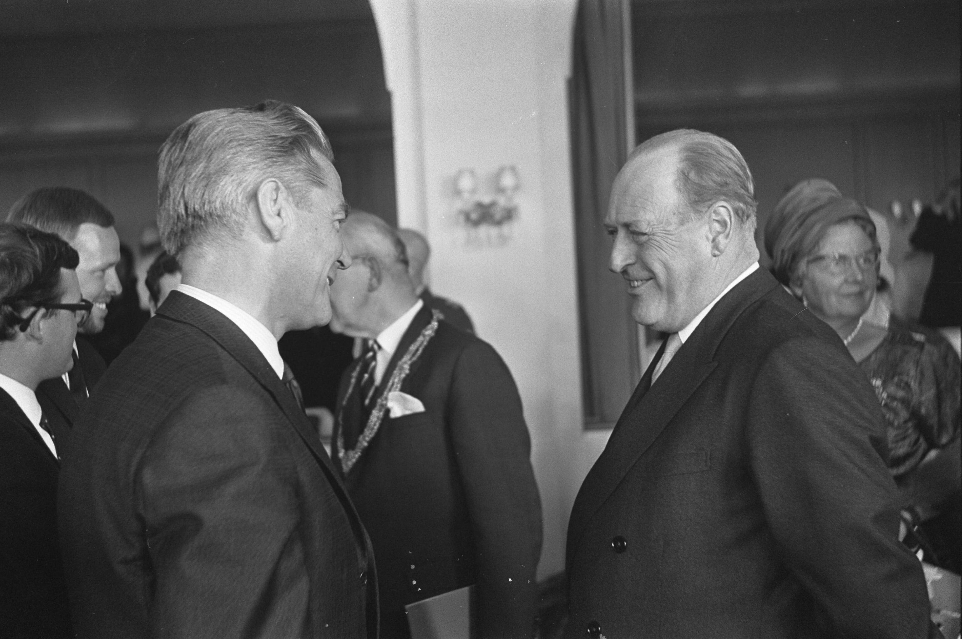 Staatsbezoek van koning Olav van Noorwegen aan Nederland — Bezoek koning Olav van Noorwegen aan Den Haag. Koning Olav in gesprek met filmer Bert Haanstra. Tussen hen in zichtbaar burgemeester Kolfschoten. Recths koningin Juliana — Foto: Nijs, Jac. de / Anefo. Licentie: Set B: Rechtenvrij / Publiek Domein. Bron: Nationaal Archief. http://hdl.handle.net/10648/aa787564-d0b4-102d-bcf8-003048976d84