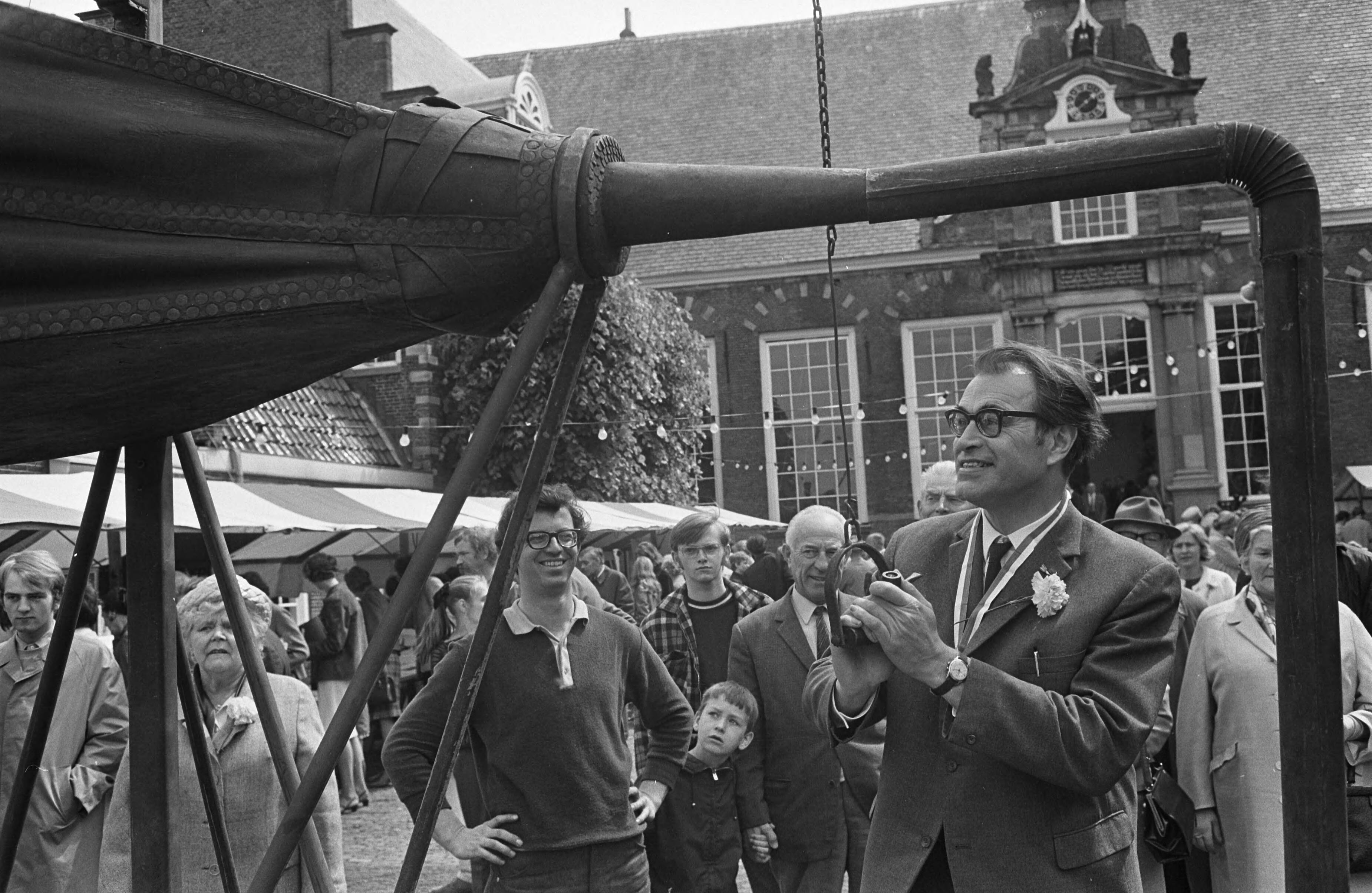Fotocollectie Anefo — Op de binnenplaats van de Kloveniers- of Stadsdoelen te Haarlem heeft de schrijver Godfried Bomans de 5e Kunstmarkt geopend. Het was ook de vijfde keer dat hij dit mocht doen. Een belangrijk deel van de opbrengst is bestemd voor het gehandicapte kind in Haarlem en omgeving. Na een korte openingsrede hielp Godfried Bomans de siersmid op de markt met het aanwakkeren van het vuur door middel van een grote blaasgalg, waarna kunstenaars aan het werk gingen — Foto: Koch, Eric / Anefo. Licentie: Set B: Rechtenvrij / Publiek Domein. Bron: Nationaal Archief. http://hdl.handle.net/10648/ab43716a-d0b4-102d-bcf8-003048976d84