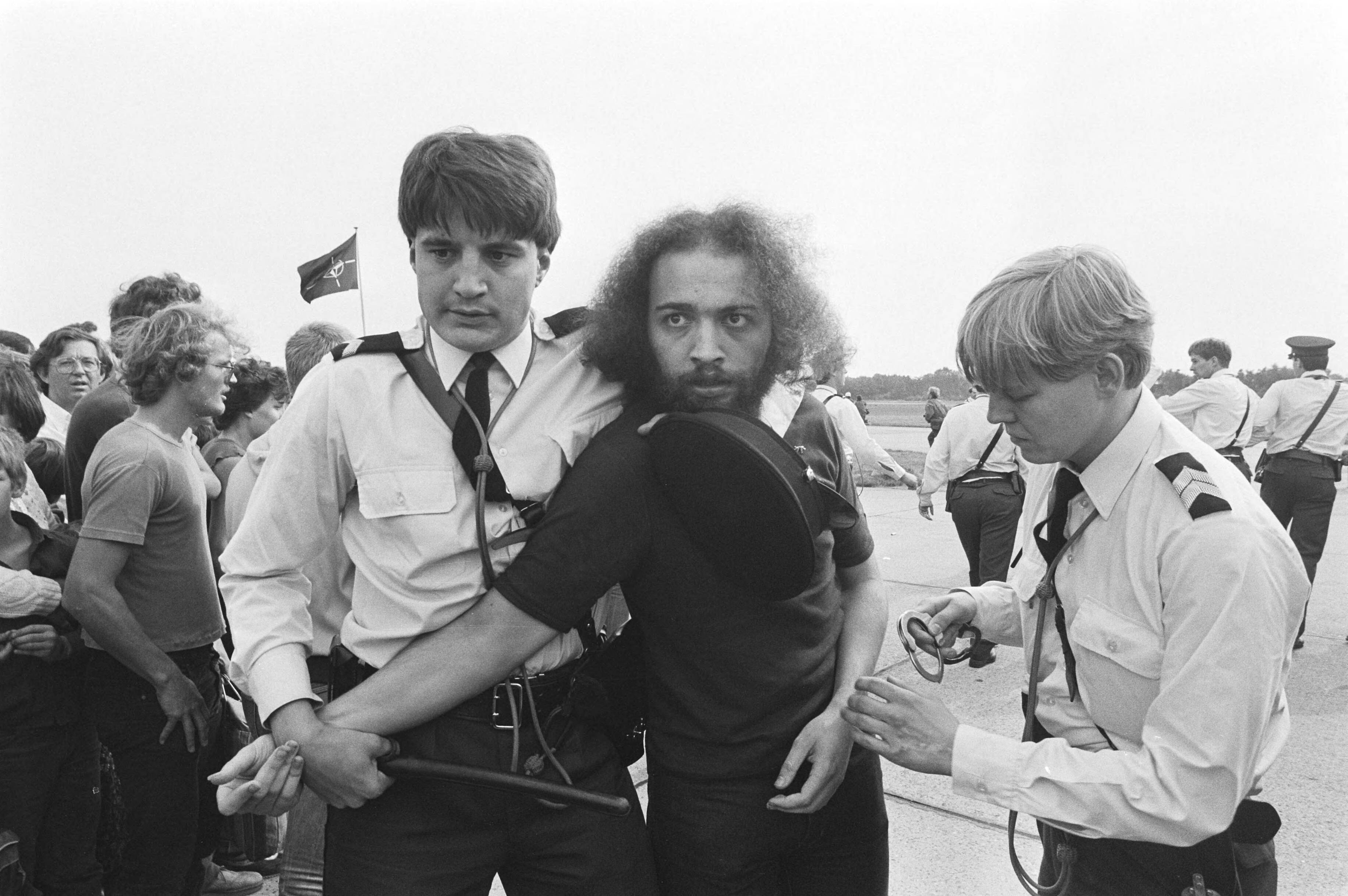 Open dag op luchtmachtbasis Soesterberg — Leden van de actiegroep "Onkruit" demonstreerden tegen de krijgsmacht op de open dag op luchtmachtbasis Soesterberg. Een groep demonstranten wordt door de Koninklijke Marechaussee weggeleid — Foto: Dijk, Hans van / Anefo. Licentie: Set B: Rechtenvrij / Publiek Domein. Bron: Nationaal Archief. http://hdl.handle.net/10648/ad0398e0-d0b4-102d-bcf8-003048976d84