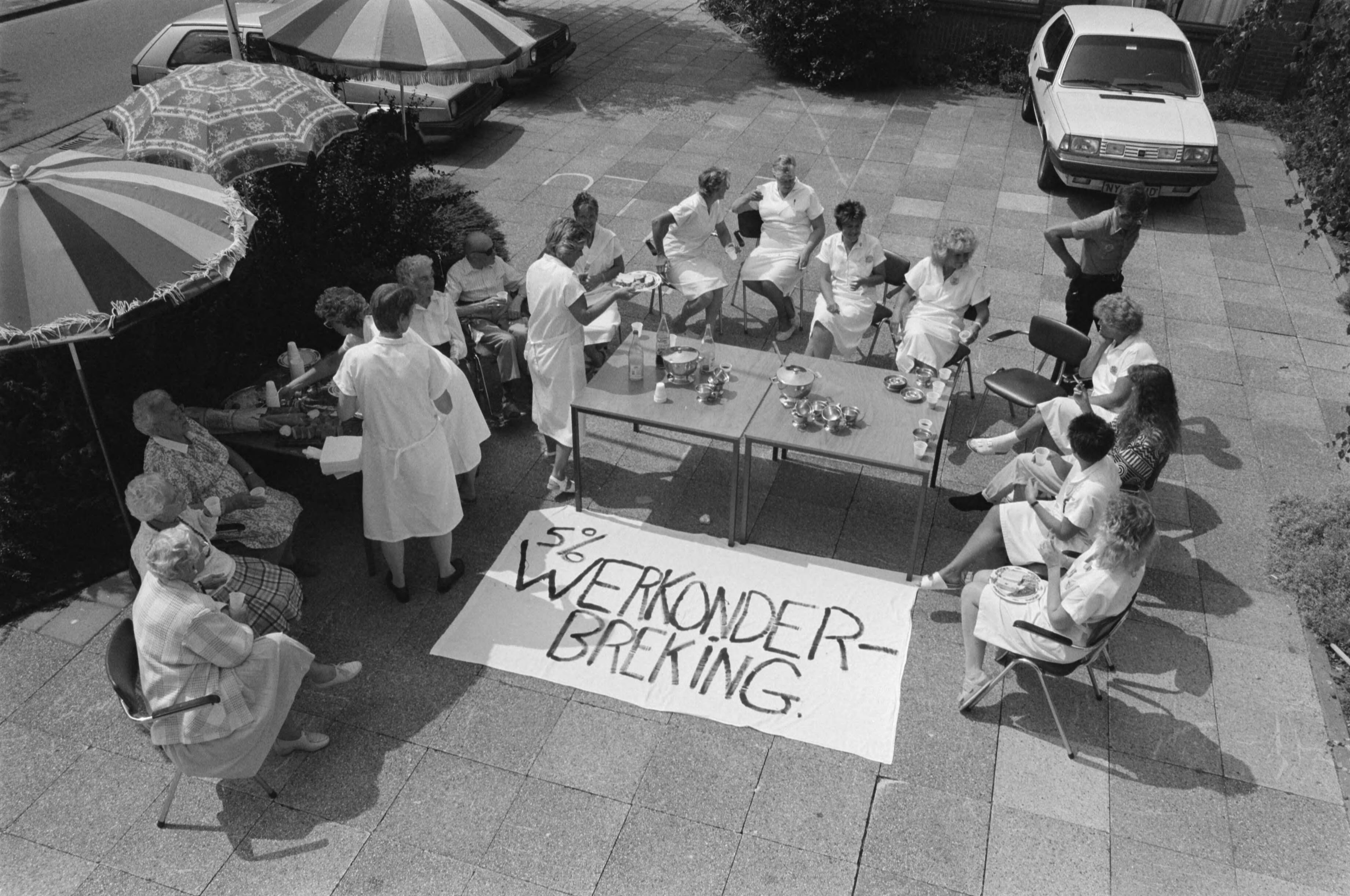 Actie in het bejaardentehuis St. Joseph in Purmerend voor 5% CAO looneis — Verplegend personeel buiten rond de tafel met een spandoek op de grond — Foto: Croes, Rob C. / Anefo. Licentie: Set B: Rechtenvrij / Publiek Domein. Bron: Nationaal Archief. http://hdl.handle.net/10648/ad73ef1e-d0b4-102d-bcf8-003048976d84