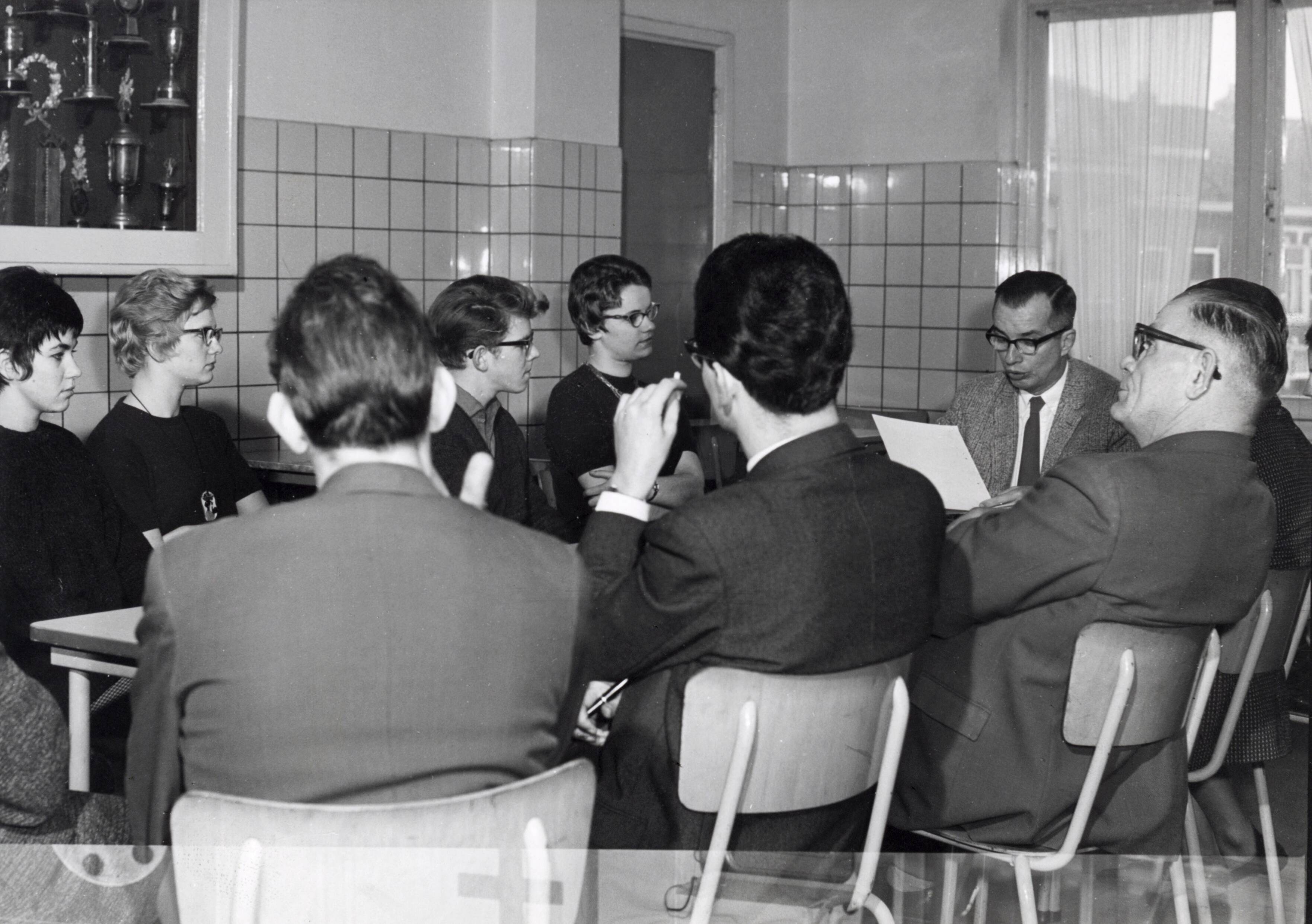 Collectie nader te bepalen — [Serie: Spaarnestad bedrijfsvoering]] [In de kantine zitten jongeren tegenover oudere heren, waarvan er een van een blad voorleest] — Foto: Fotograaf Onbekend / Fotobureau onbekend / Spaarnestad Photo (05887). Licentie: Set B: Rechtenvrij / Publiek Domein. Bron: Nationaal Archief. http://hdl.handle.net/10648/35429da6-414b-a382-e063-6df0900a6686