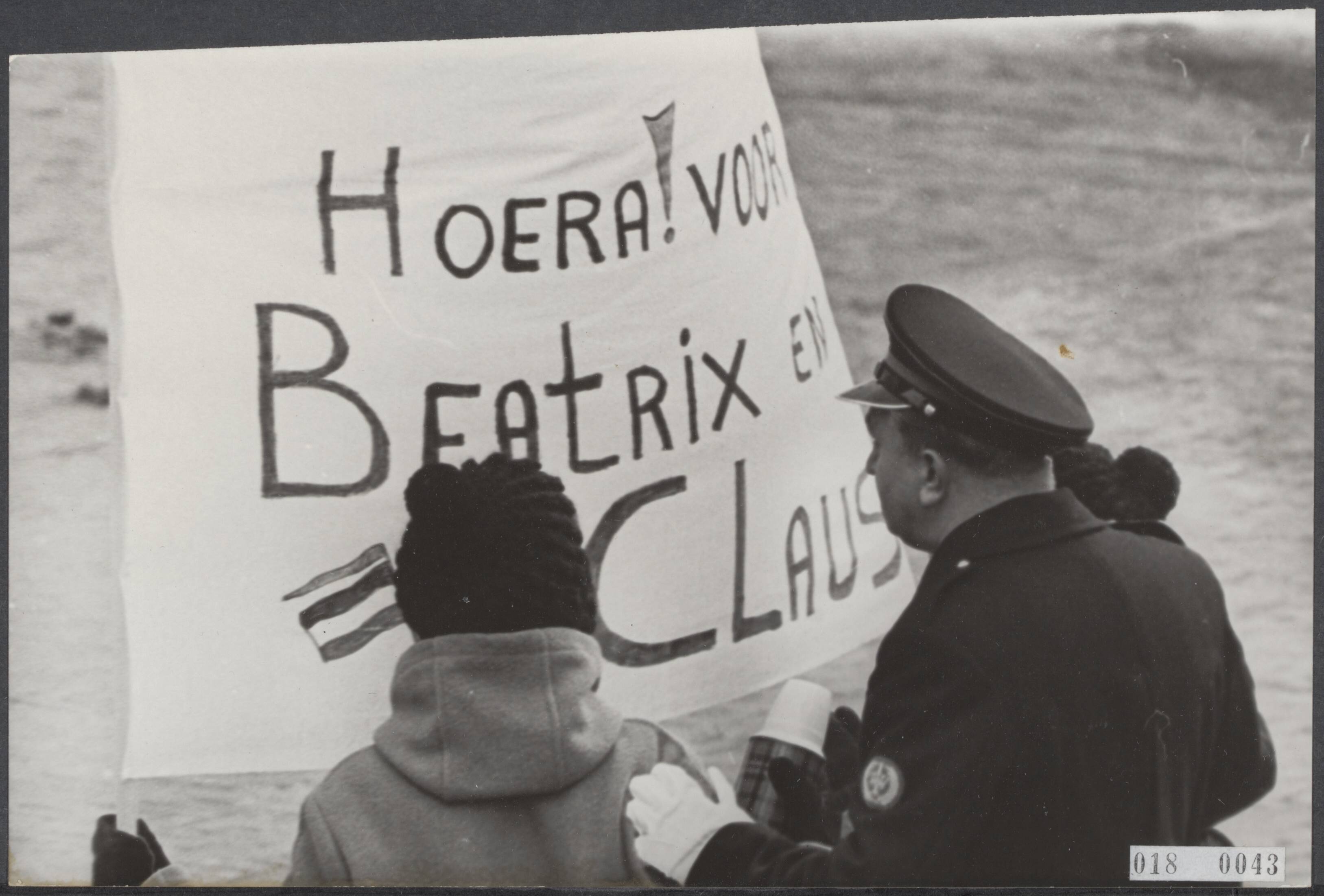 Fotocollectie Elsevier Binnenland — Prinses Beatrix en Claus von Amsberg zijn in Baarn in ondertrouw gegaan. Twee jongeren met een spandoek met een geschreven felicitatie worden door een politieagent achter de afzetting gebracht — Foto: Fotograaf Onbekend / Anefo. Licentie: Set B: Rechtenvrij / Publiek Domein. Bron: Nationaal Archief. http://hdl.handle.net/10648/adc108ee-d0b4-102d-bcf8-003048976d84
