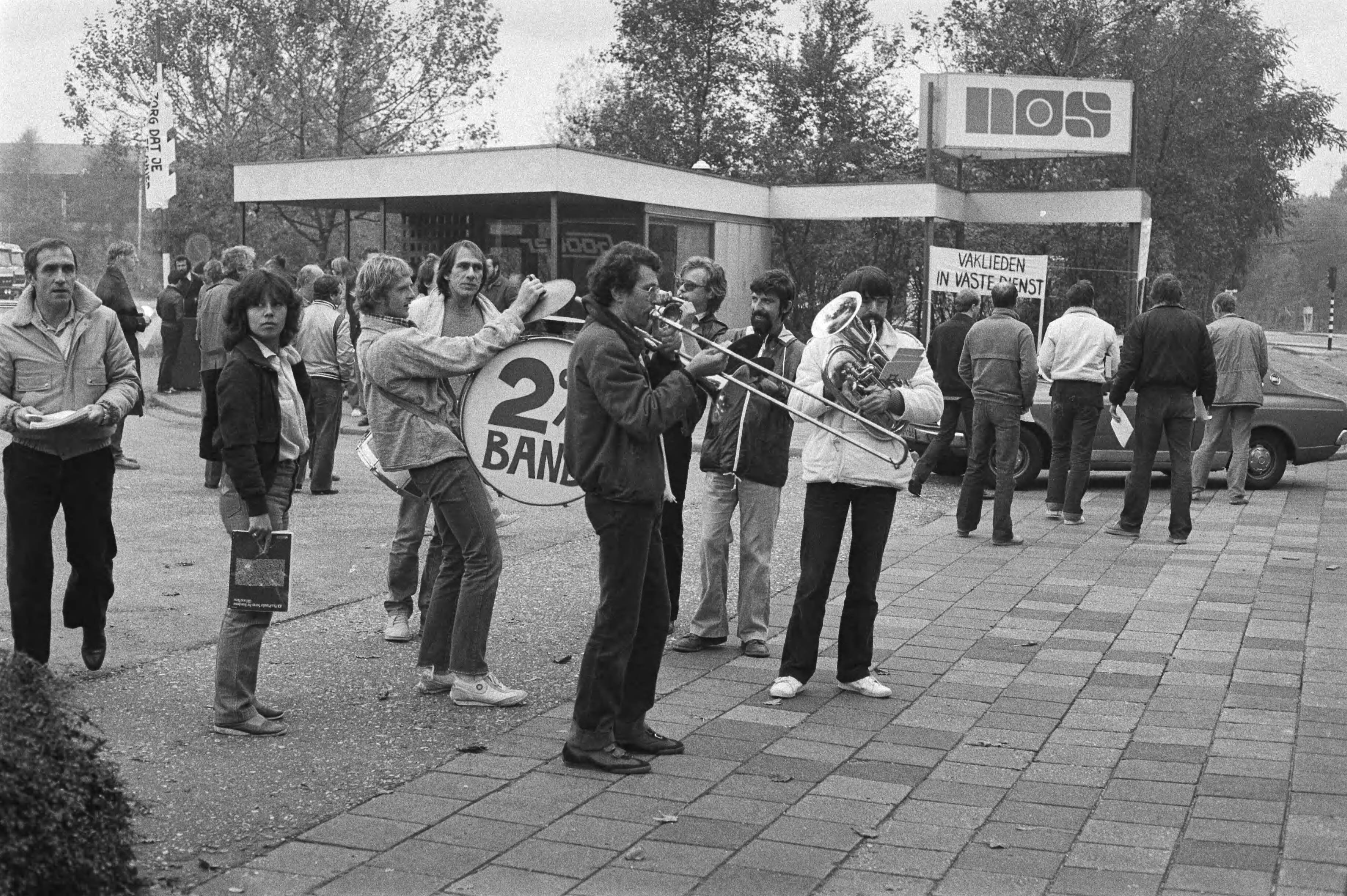 Staking bij de NOS in Hilversum — Postende omroepmedewerkers maken muziek voor de toegangspoort van de NOS in Hilversum — Foto: Antonisse, Marcel / Anefo. Licentie: Set B: Rechtenvrij / Publiek Domein. Bron: Nationaal Archief. http://hdl.handle.net/10648/ad065fd0-d0b4-102d-bcf8-003048976d84