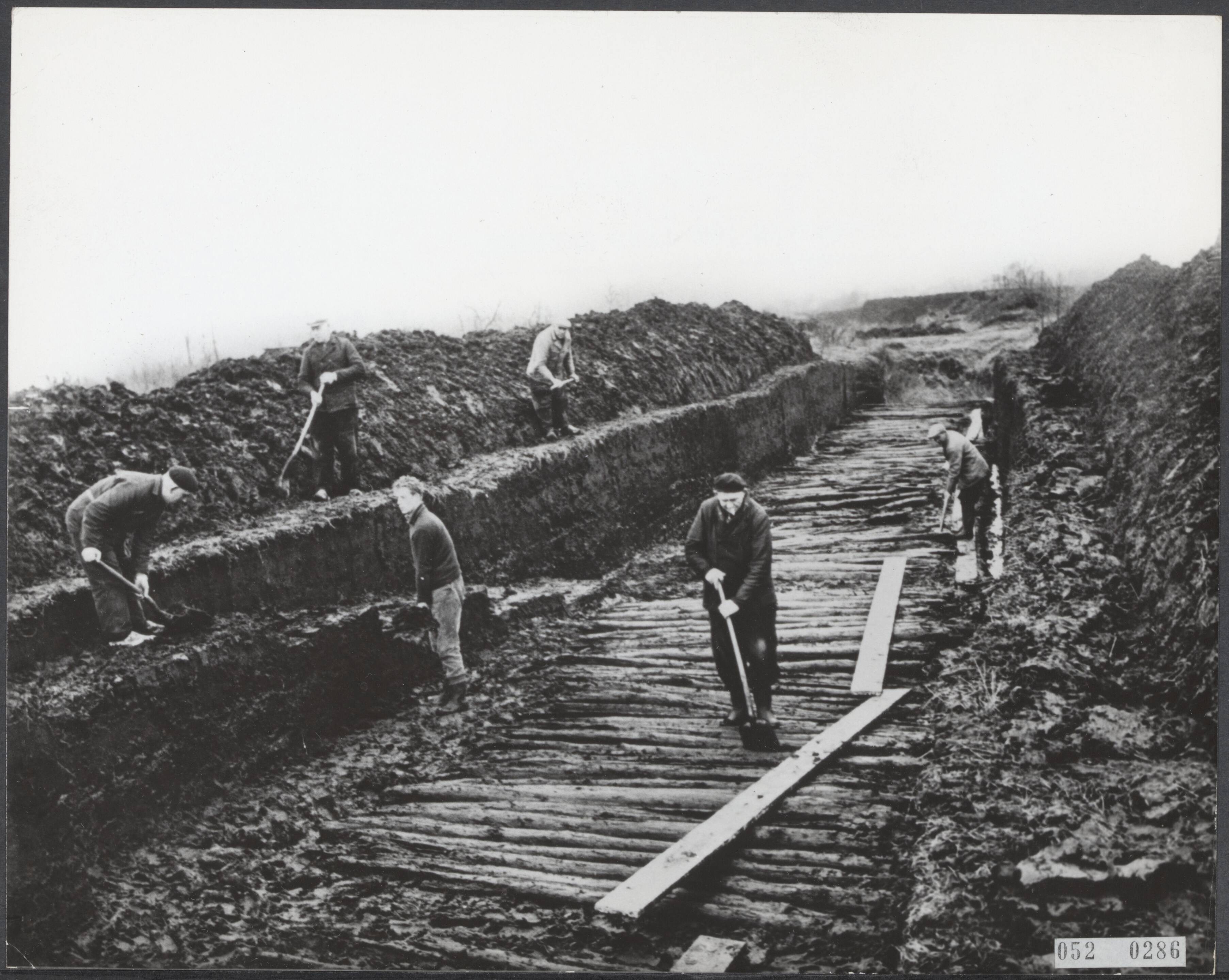 Fotocollectie Elsevier Binnenland — Ten zuidoosten van Emmen is een veenbrug ontdekt. Een houten weg van balken en stammetjes ongeveer 3 meter breed, daterende van 2000 jaar vóór Christus. Nog dit jaar zal 50 meter van de brug worden opgegraven o.l.v. drs. W Casparie van het Biologisch Archeologisch Instituut te Grongen. Als gast is hierbij aanwezig dr. MacAndrews uit Minnesota (Amerika) — Foto: Kroon, Ron / Anefo. Licentie: Set B: Rechtenvrij / Publiek Domein. Bron: Nationaal Archief. http://hdl.handle.net/10648/ae057dee-d0b4-102d-bcf8-003048976d84