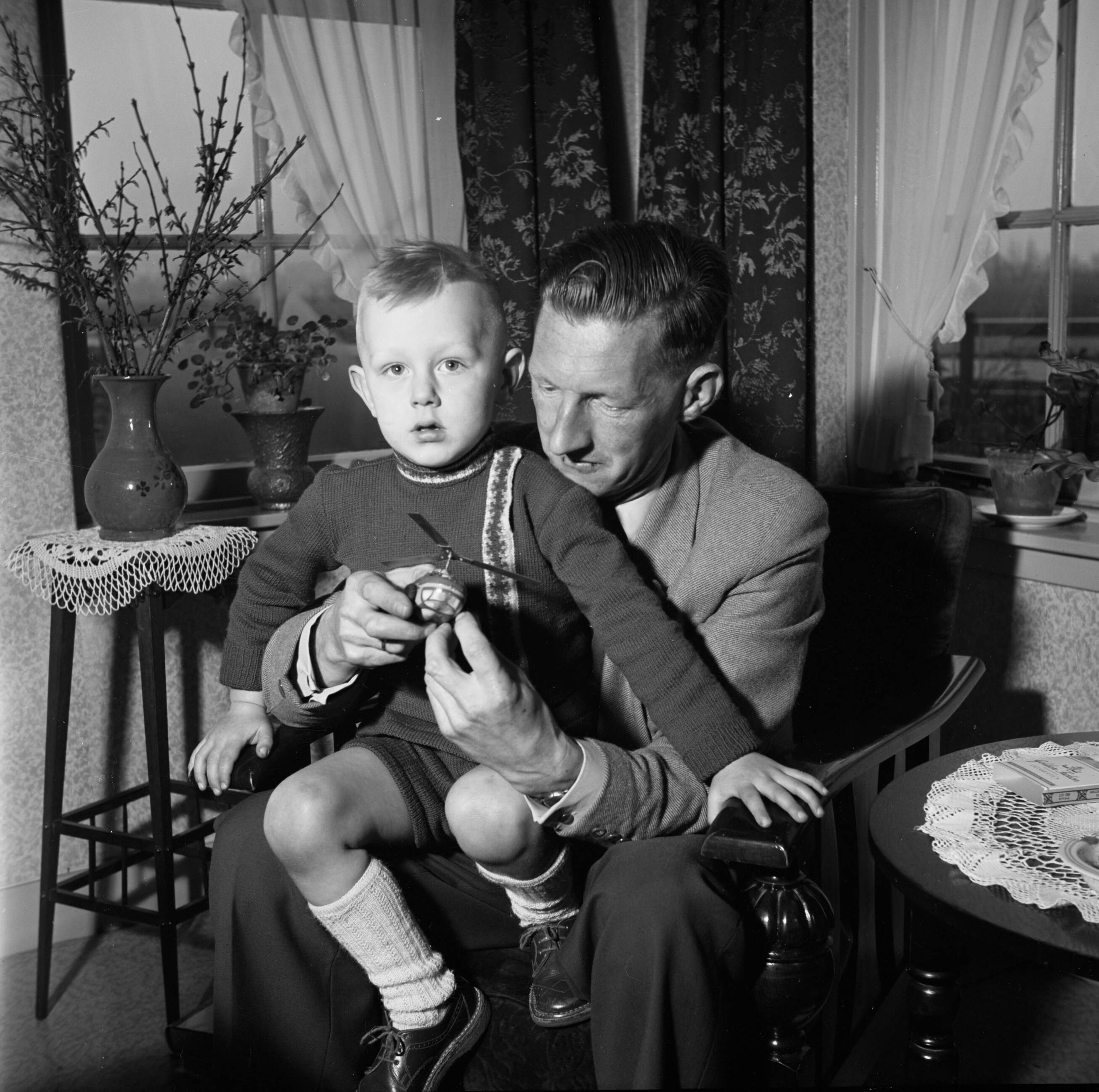 Fotocollectie Van de Poll — Gerard met zijn zoontje Gerrie op schoot terwijl hij een speelgoedhelikopter in de hand houdt. Links een bloementafeltje met een vaas met uitbottende takken — Foto: Poll, Willem van de. Licentie: Set B: Rechtenvrij / Publiek Domein. Bron: Nationaal Archief. http://hdl.handle.net/10648/aeccd2fe-d0b4-102d-bcf8-003048976d84