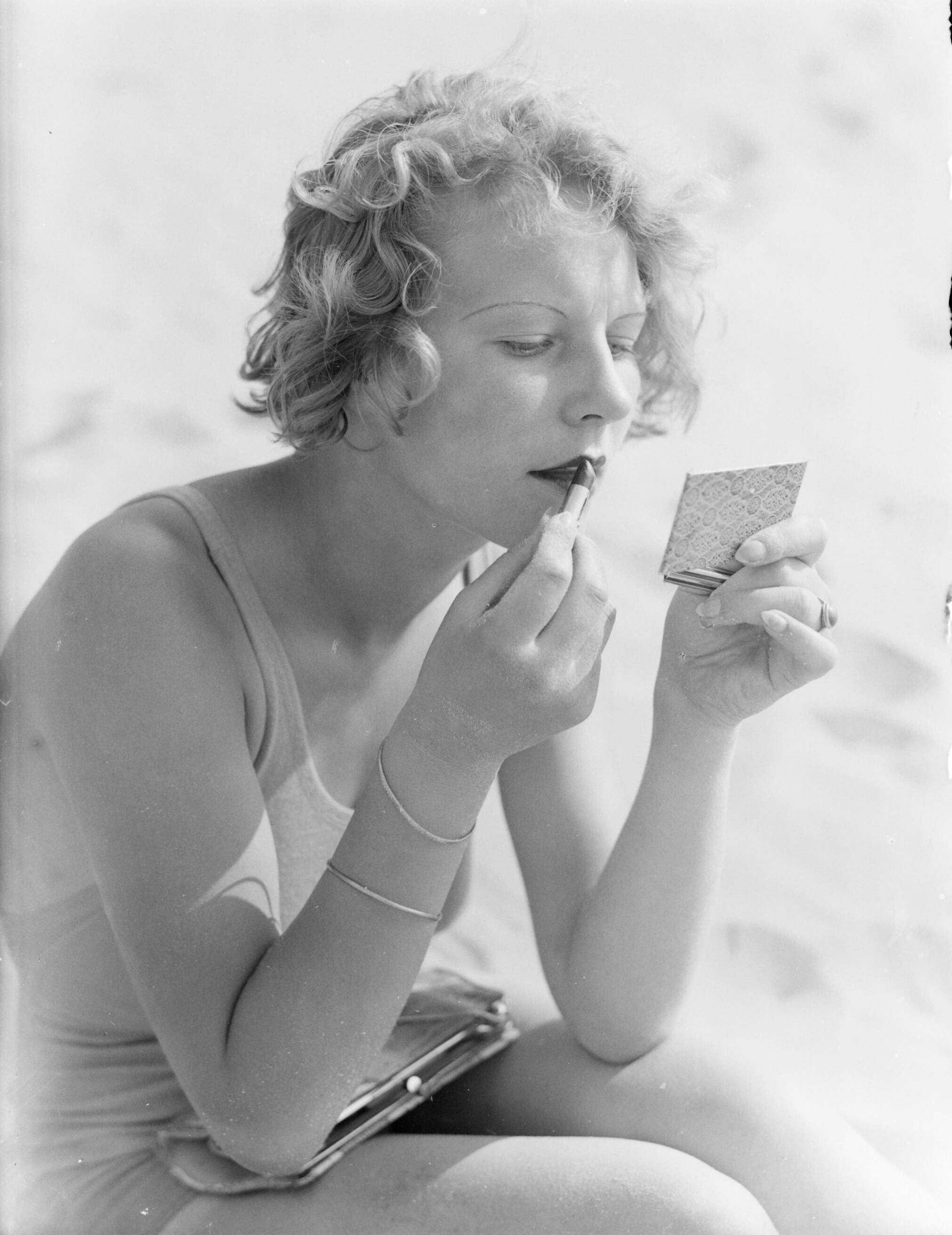 Reportage fotomodel — Model Eva Waldschmidt zit in het zand en stift haar lippen — Foto: Poll, Willem van de. Licentie: Set B: Rechtenvrij / Publiek Domein. Bron: Nationaal Archief. http://hdl.handle.net/10648/aea7f966-d0b4-102d-bcf8-003048976d84