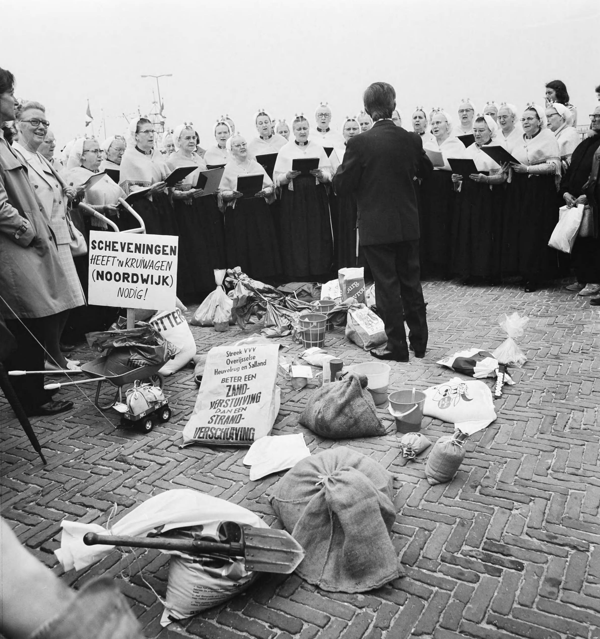 Fotocollectie Anefo — VVV-directeuren brengen zak zand naar jubilerende VVV in Scheveningen; zakken zand op boulevard met zangkoor — Foto: Mieremet, Rob / Anefo. Licentie: Set B: Rechtenvrij / Publiek Domein. Bron: Nationaal Archief. http://hdl.handle.net/10648/ac4ae098-d0b4-102d-bcf8-003048976d84