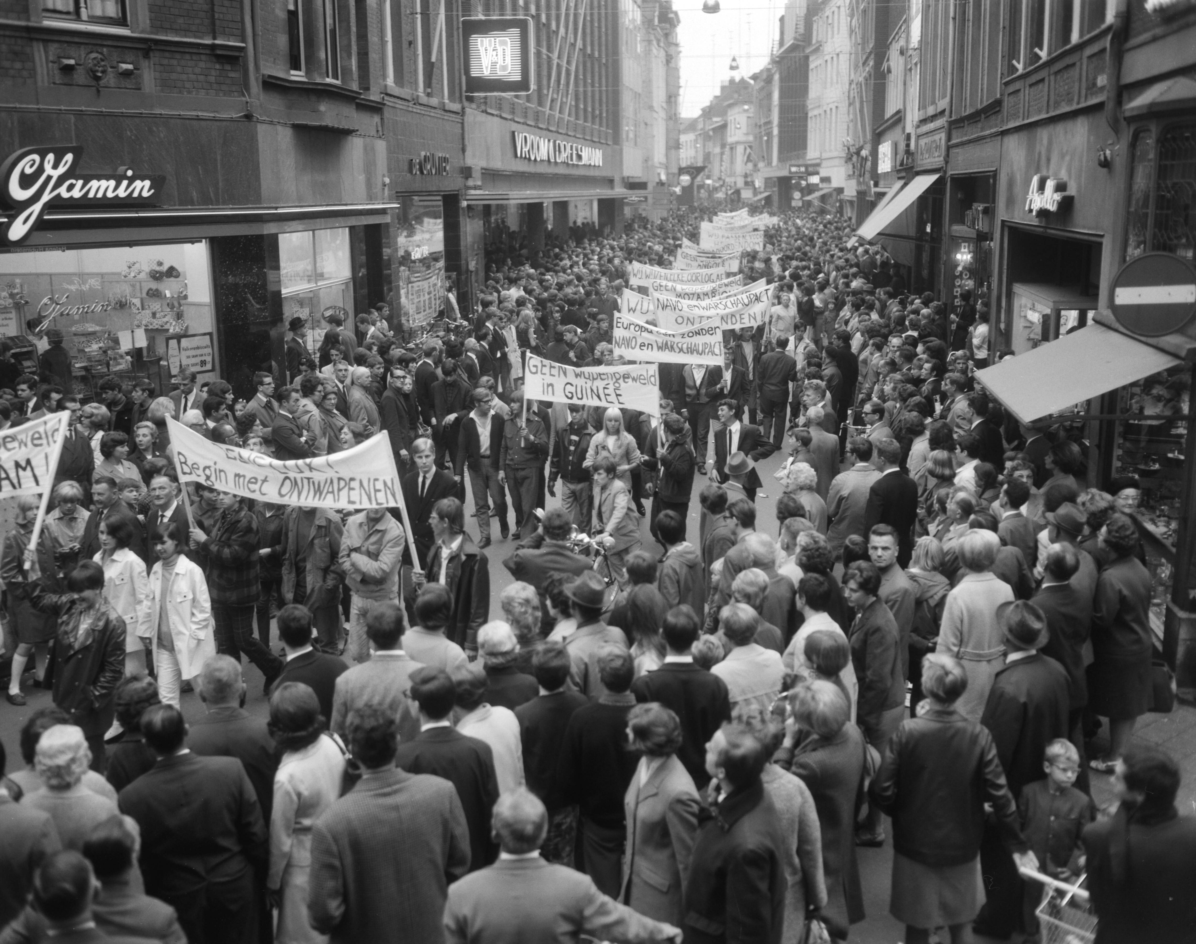 Anti NAVO-demonstratie in Maastricht — Demonstranten met spandoeken in de Grote Straat — Foto: Bilsen, Joop van / Anefo. Licentie: Set B: Rechtenvrij / Publiek Domein. Bron: Nationaal Archief. http://hdl.handle.net/10648/aafde320-d0b4-102d-bcf8-003048976d84