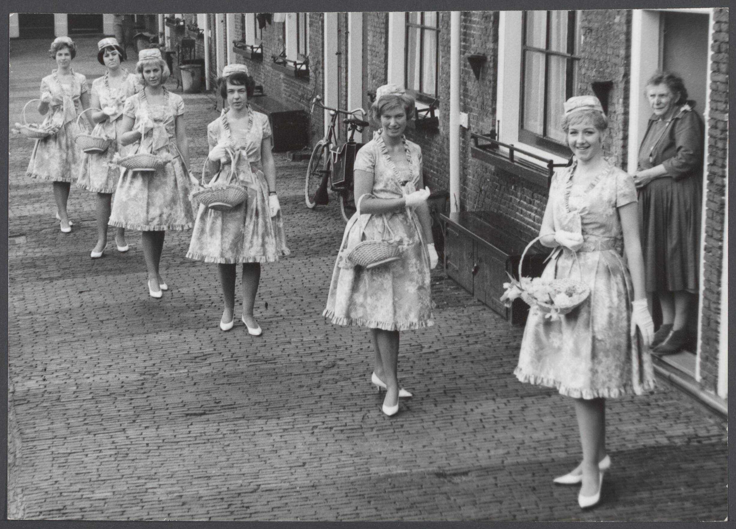 Bloemenmeisjes III — De Haarlemse Bloemenmeisjes werden aan de pers gepresenteerd in hun nieuwe Boussac kantoene jurkjes met groen vilten jassen. Zij zullen, 24 in getal, vanaf 21 maart wanneer de lente begint als gastvrouwen voor Haarlem optreden. Enkele meisjes waaonder Carla, Adri, Inez, Iris en Pien op een der straatjes in het Proveniershofje in Haarlem — Foto: Nijs, Jac. de / Anefo. Licentie: Set B: Rechtenvrij / Publiek Domein. Bron: Nationaal Archief. http://hdl.handle.net/10648/6b5cb2f2-9e83-7c69-4b08-fd58c554a7ff