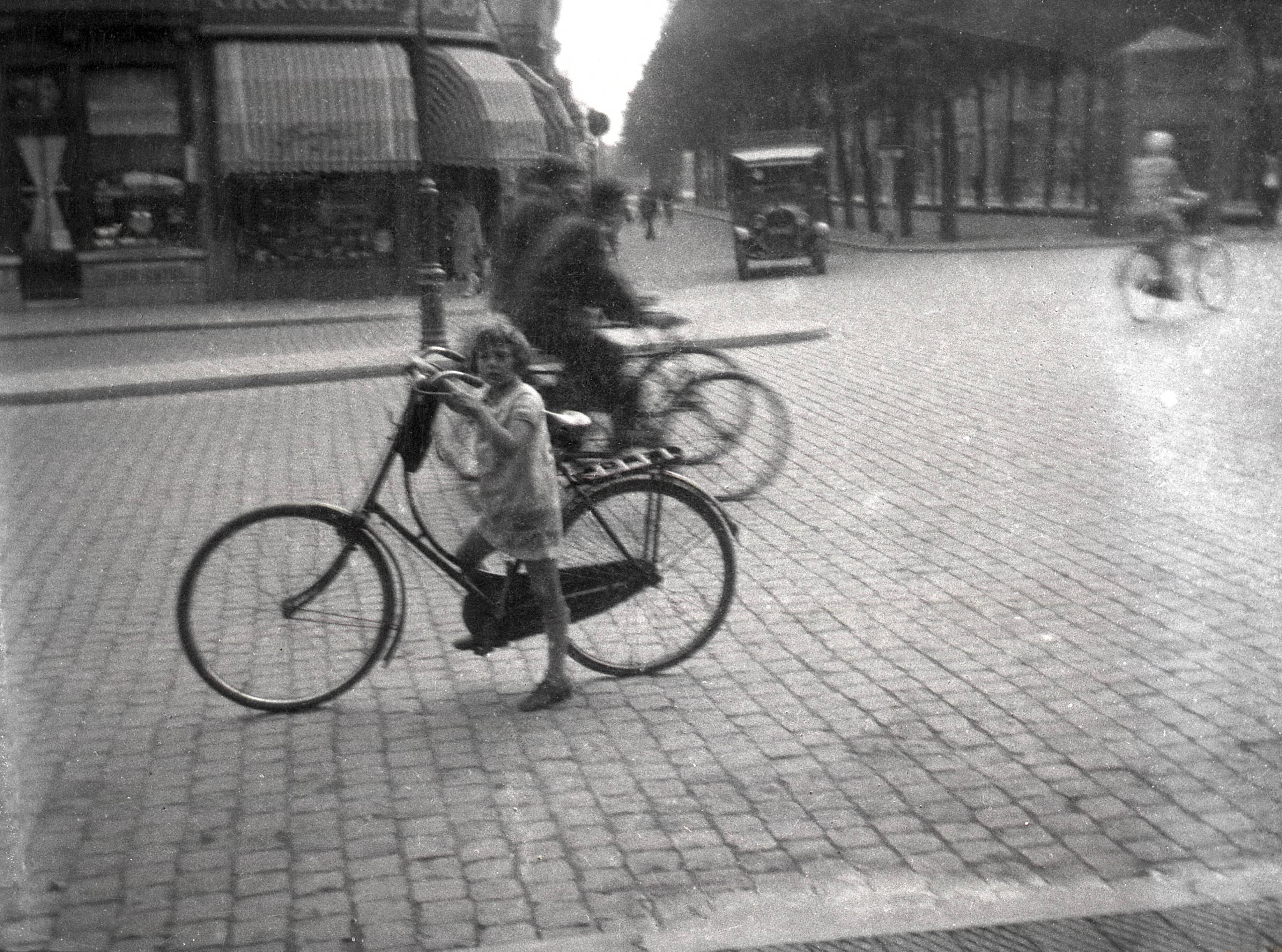 Portretten, Verkeer, "Topografie, plaatsen op naam" — Fietsen in Nederland. Een meisje staat met een damesfiets (opoefiets)op het met kinderhoofdjes geplaveide Van Coothplein te Breda. Op de achtergrond fietsers en een automobiel (vrachtauto). Omstreeks 1928. De foto is afkomstig uit de collectie van portretfotograaf bekend onder de naam 'Atelier Anton Henning'. — Foto: Fotograaf onbekend / Fotobureau onbekend / Rechtenvrij (05889). Licentie: Set B: Rechtenvrij / Publiek Domein. Bron: Nationaal Archief. http://hdl.handle.net/10648/35429da6-64ba-a382-e063-6df0900a6686