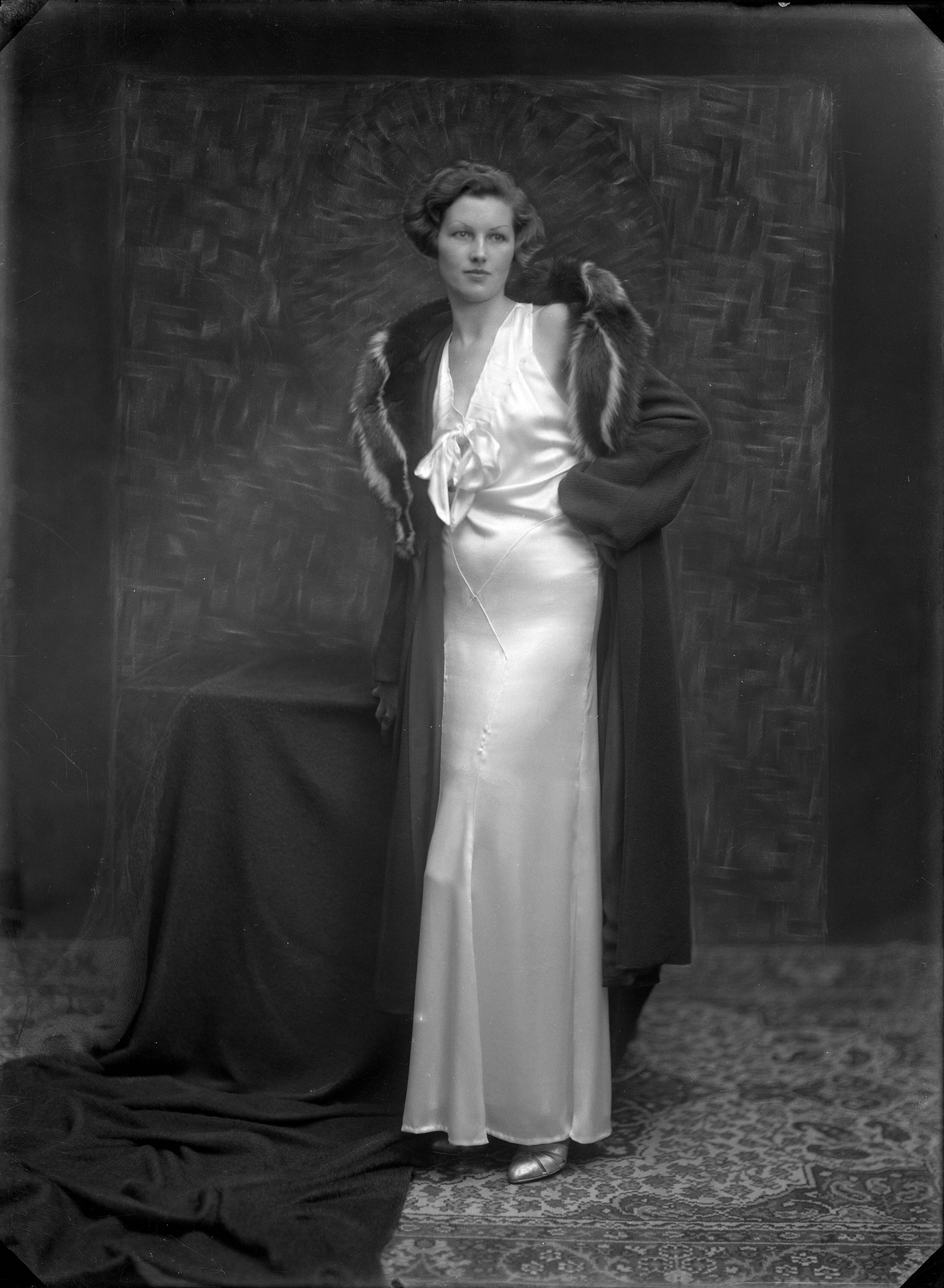 Berg, van den (BERG 050) — Studio-opname van een vrouw in een lange witte satijnen jurk met een lange jas met bontkraag erover. Haarlem, Nederland 1918-1930 — Foto: Berg, Th.C.J. van den / Spaarnestad Photo (05887). Licentie: Set B: Rechtenvrij / Publiek Domein. Bron: Nationaal Archief. http://hdl.handle.net/10648/35429da7-c554-a382-e063-6df0900a6686