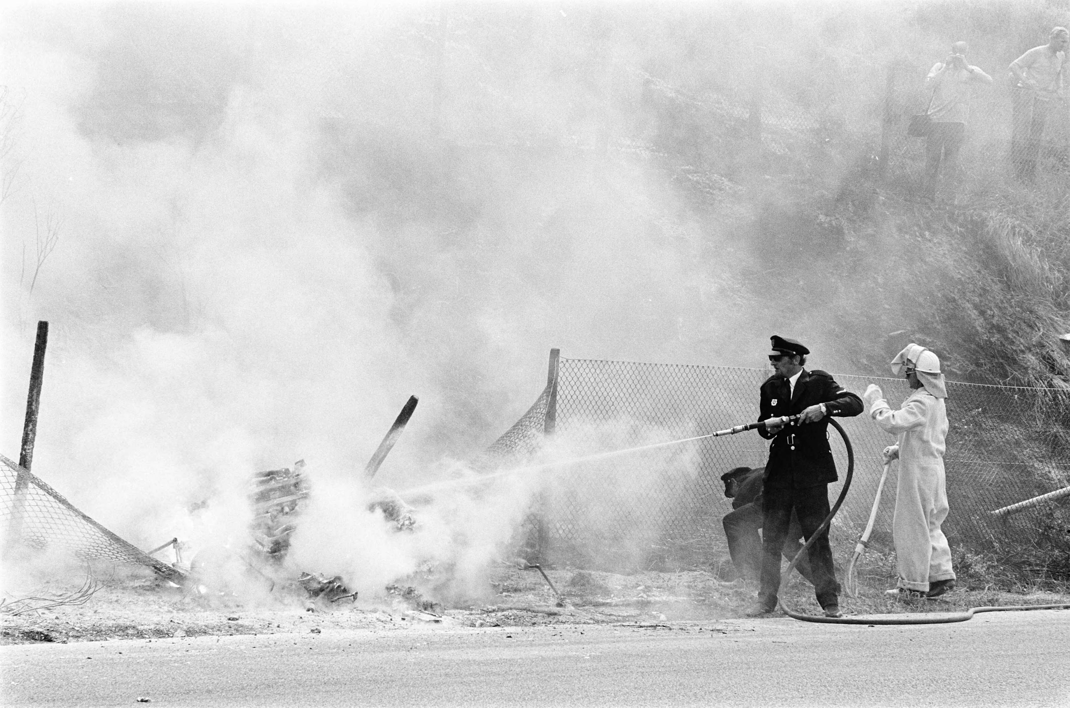 Grand Prix Zandvoort 1970 — Grand Prix 1970 van Nederland voor Formule I wagens , Zandvoort; auto van Piers Courage gecrasht en in brand gevlogen. Een politieman helpt bij het blussen — Foto: Evers, Joost / Anefo. Licentie: Set B: Rechtenvrij / Publiek Domein. Bron: Nationaal Archief. http://hdl.handle.net/10648/ab9a483c-d0b4-102d-bcf8-003048976d84