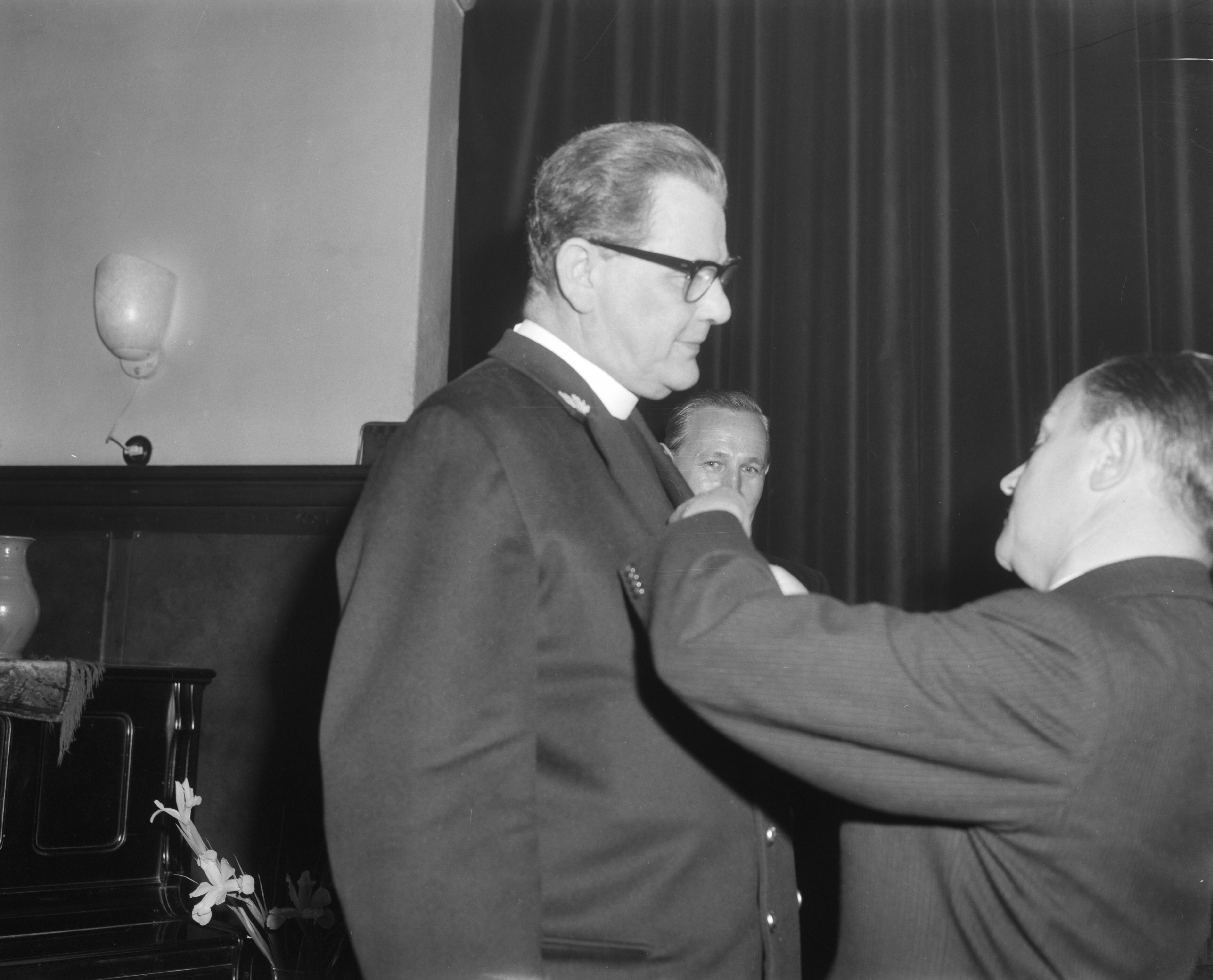 Afscheidsreceptie van Hoofd Vloot Aalmoezenier kapitein ter Zee Mgr. J.F.M. de Sain in RK militair tehuis Den Haag — Afscheidsreceptie van de Hoofdvlootaalmoezenier kapitein-ter-zee mgr. J.F.M. de Sain in het R.K. militair tehuis in Den Haag. Mgr. De Sain krijgt een koninklijke onderscheiding opgespeld door staatssecretaris De Jong — Foto: Bilsen, Joop van / Anefo. Licentie: Set B: Rechtenvrij / Publiek Domein. Bron: Nationaal Archief. http://hdl.handle.net/10648/aa3b3956-d0b4-102d-bcf8-003048976d84