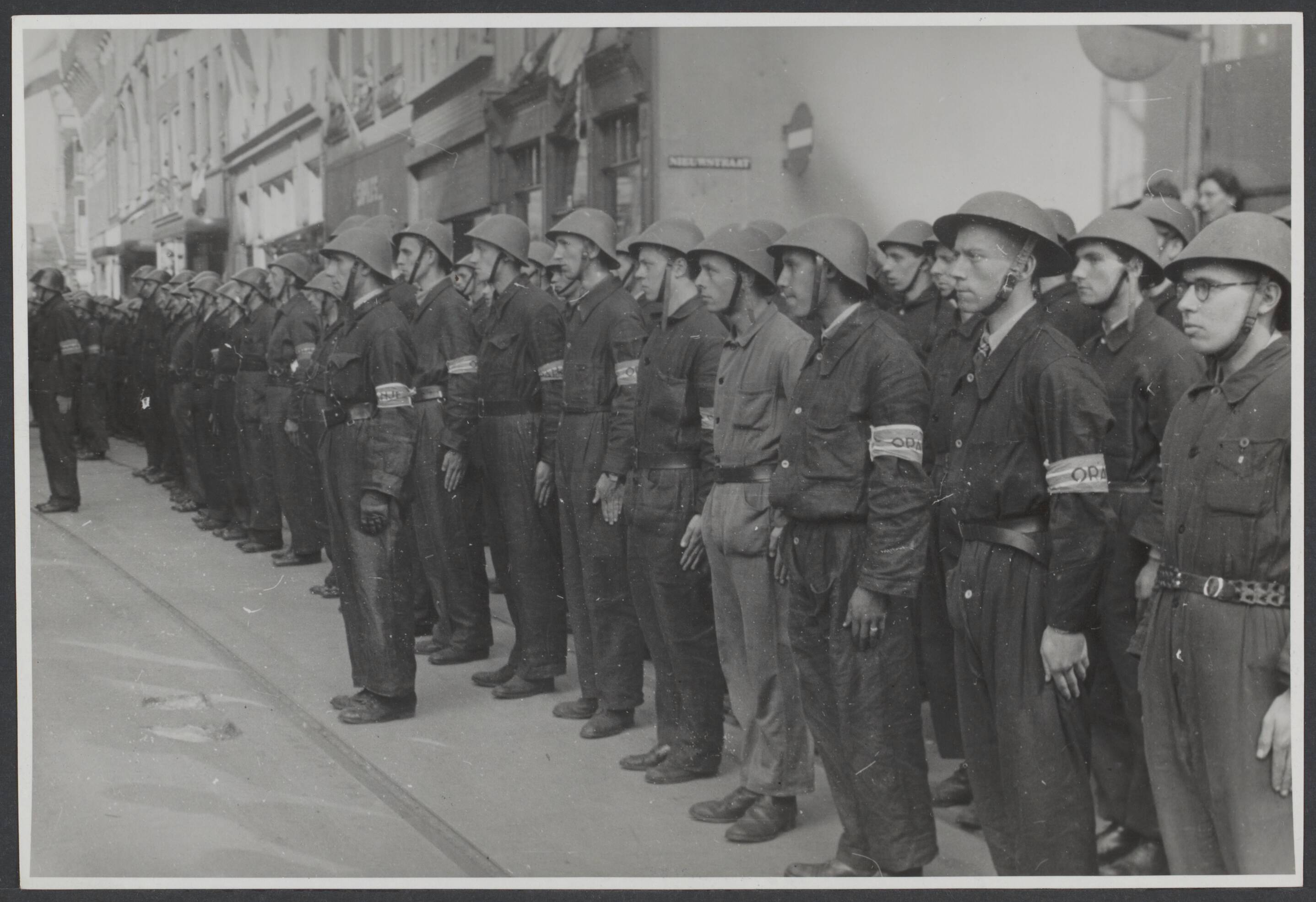 Bevrijding 's Gravenhage (Den Haag) — De Nederlandse Binnenlandse Strijdkrachten (BS) in blauwe overall en oranje band om de arm in het gelid voor het stadhuis, tijdens de intocht der Canadezen te Den Haag — Foto: Wal, J.Th. A. van der / Spaarnestad. Licentie: Set B: Rechtenvrij / Publiek Domein. Bron: Nationaal Archief. http://hdl.handle.net/10648/2b1fbb05-4928-edae-232c-d0730297118e