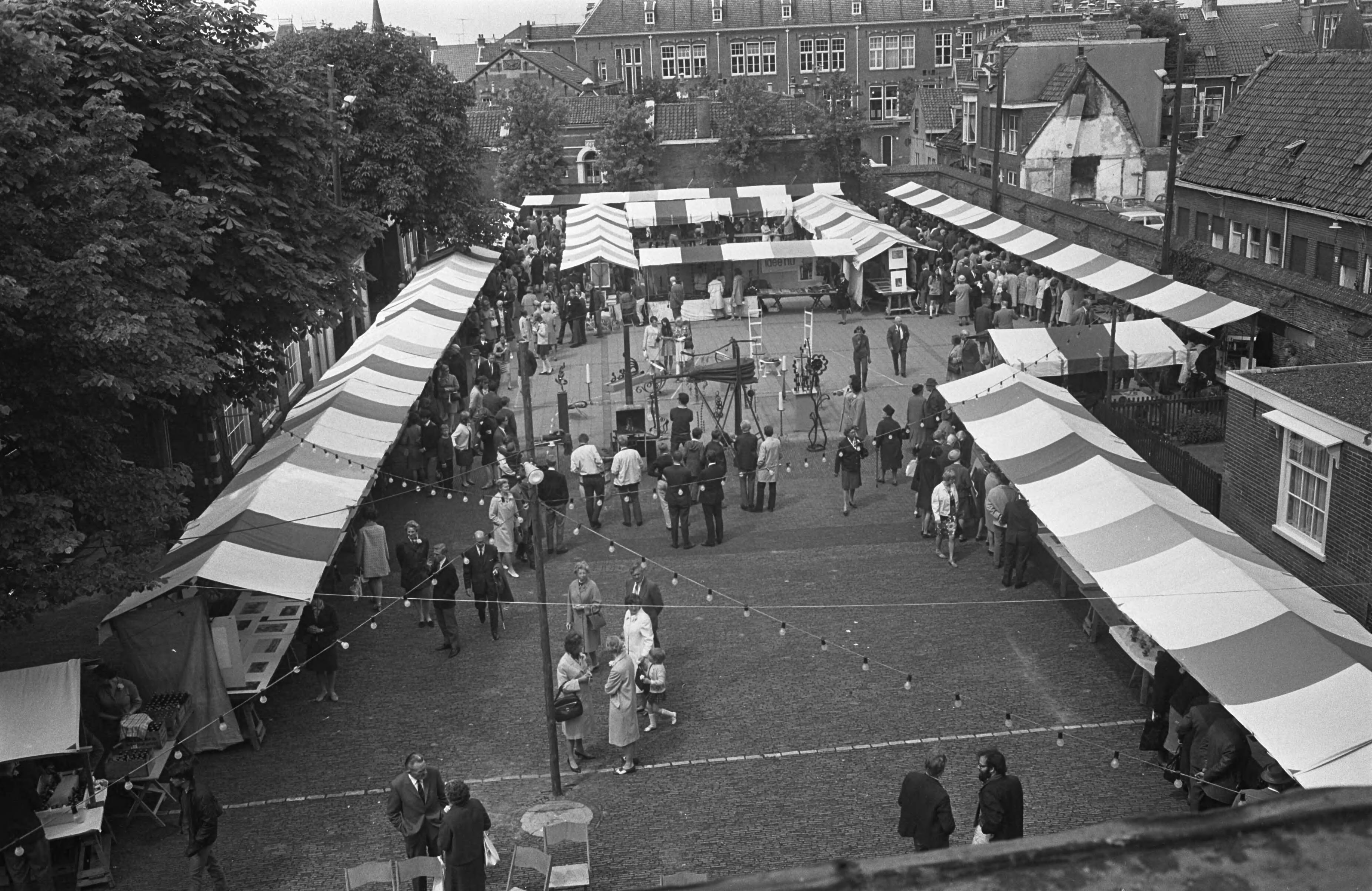 Fotocollectie Anefo — Op de binnenplaats van de Kloveniers- of Stadsdoelen te Haarlem heeft de schrijver Godfried Bomans de 5e Kunstmarkt geopend. Het was ook de vijfde keer dat hij dit mocht doen. Een belangrijk deel van de opbrengst is bestemd voor het gehandicapte kind in Haarlem en omgeving. Na een korte openingsrede hielp Godfried Bomans de siersmid op de markt met het aanwakkeren van het vuur door middel van een grote blaasgalg, waarna kunstenaars aan het werk gingen — Foto: Koch, Eric / Anefo. Licentie: Set B: Rechtenvrij / Publiek Domein. Bron: Nationaal Archief. http://hdl.handle.net/10648/ab43725a-d0b4-102d-bcf8-003048976d84