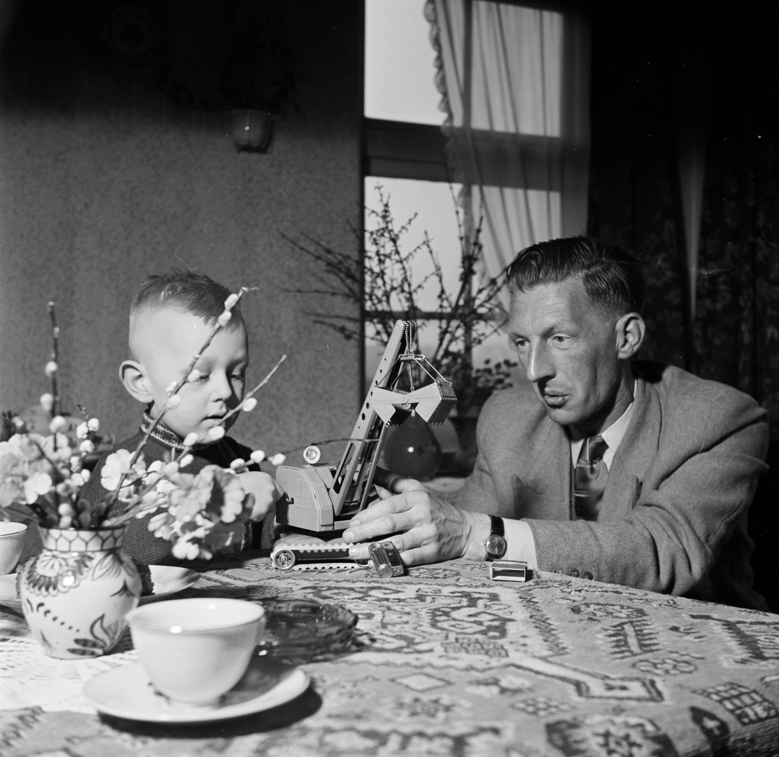 Fotocollectie Van de Poll — Gerard met zijn vierjarig zoontje Gerrie aan tafel met een speelgoedhijskraan — Foto: Poll, Willem van de. Licentie: Set B: Rechtenvrij / Publiek Domein. Bron: Nationaal Archief. http://hdl.handle.net/10648/aecccf3e-d0b4-102d-bcf8-003048976d84