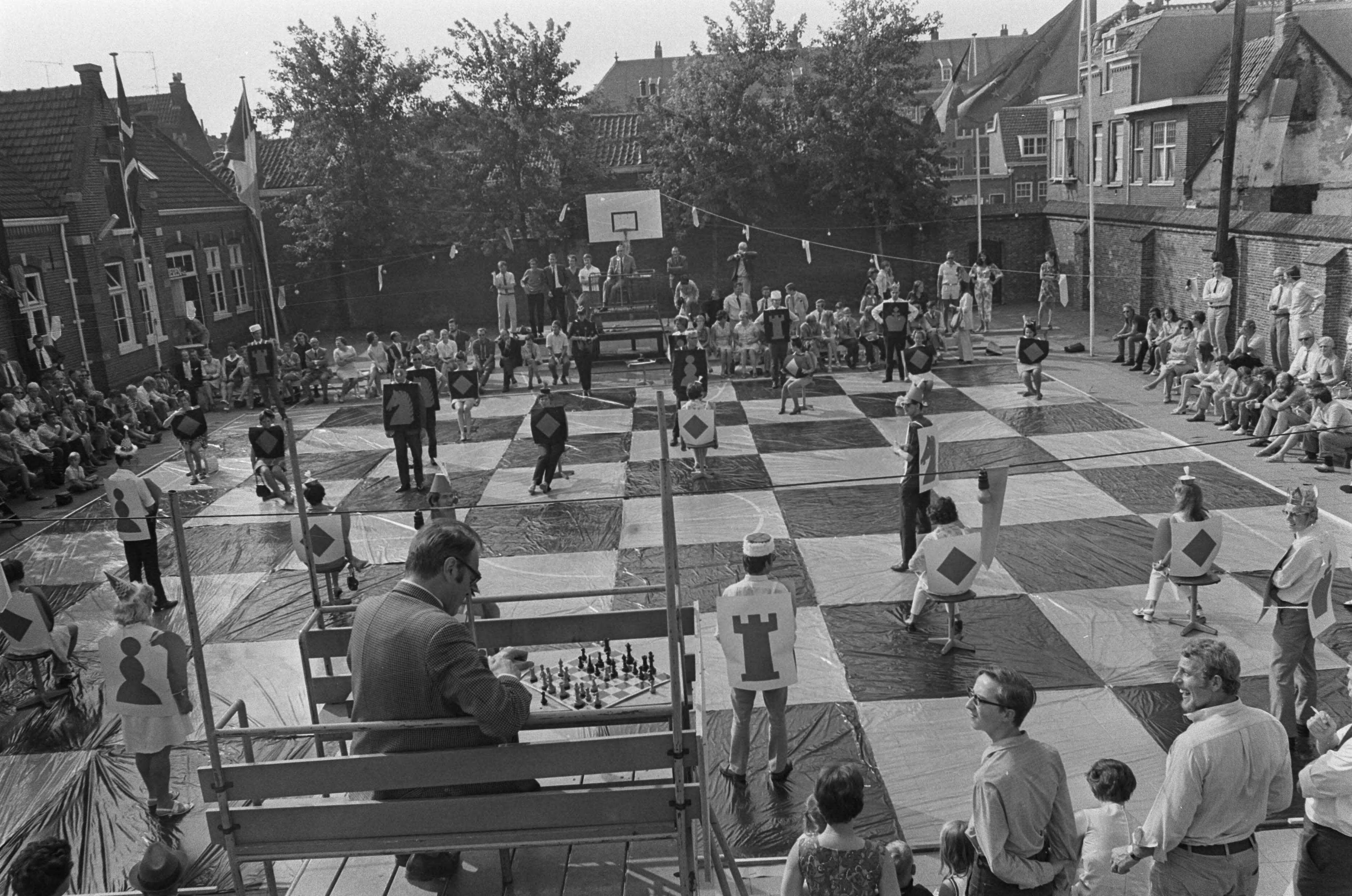 Fotocollectie Anefo — Godfried Bomans en dr. Max Euwe spelen schaakspel met levende stukken, Haarlem; overzicht, met op voorgrond G. Bomans — Foto: Mieremet, Rob / Anefo. Licentie: Set B: Rechtenvrij / Publiek Domein. Bron: Nationaal Archief. http://hdl.handle.net/10648/aba12e54-d0b4-102d-bcf8-003048976d84
