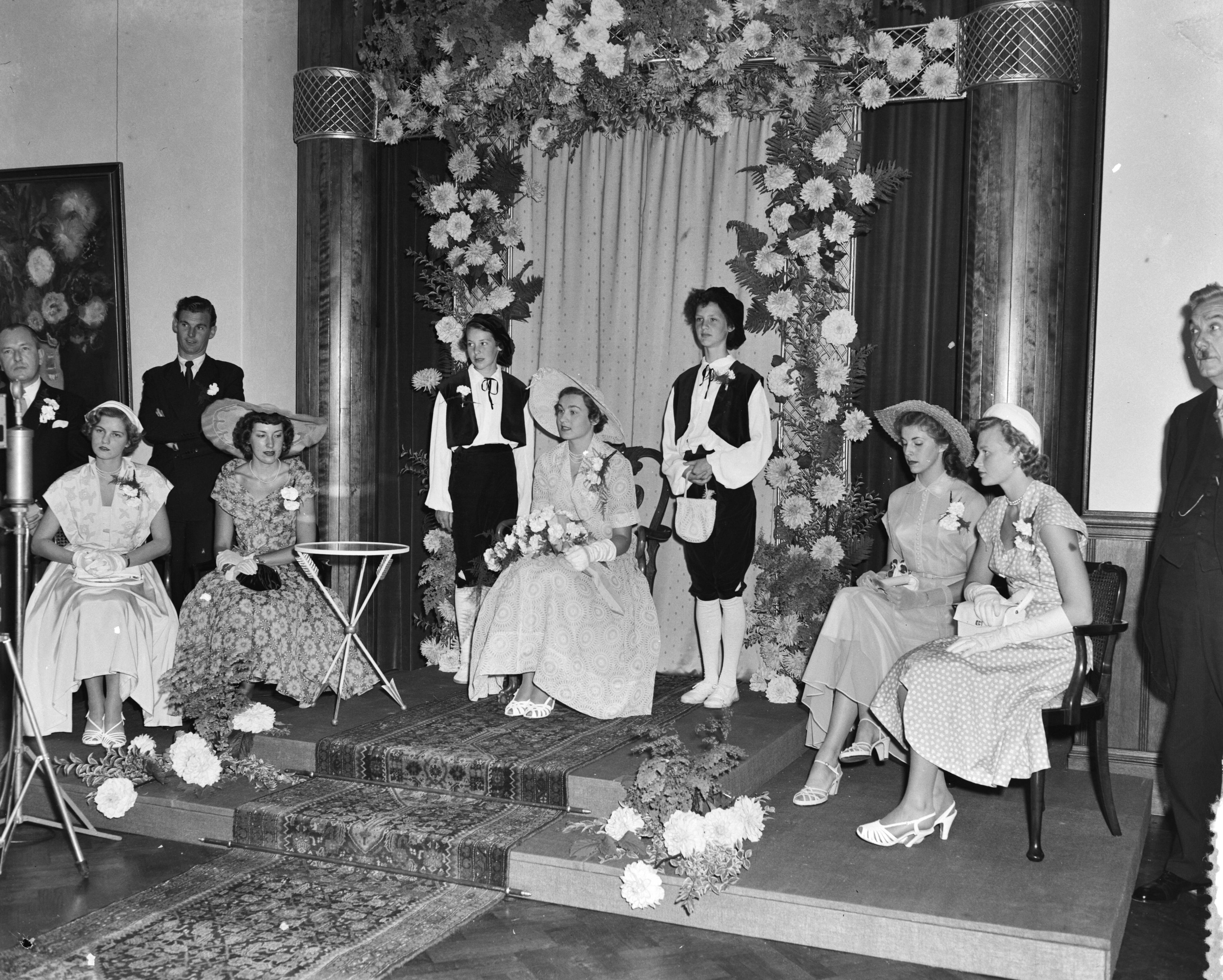 Intocht Bloemenkoningin van Den Haag — Intocht Bloemenkoningin van Den Haag. Zij zit in de oude raadszaal geflankeerd door pages en hofdames — Foto: Pot, Harry / Anefo. Licentie: Set B: Rechtenvrij / Publiek Domein. Bron: Nationaal Archief. http://hdl.handle.net/10648/a8edcc62-d0b4-102d-bcf8-003048976d84