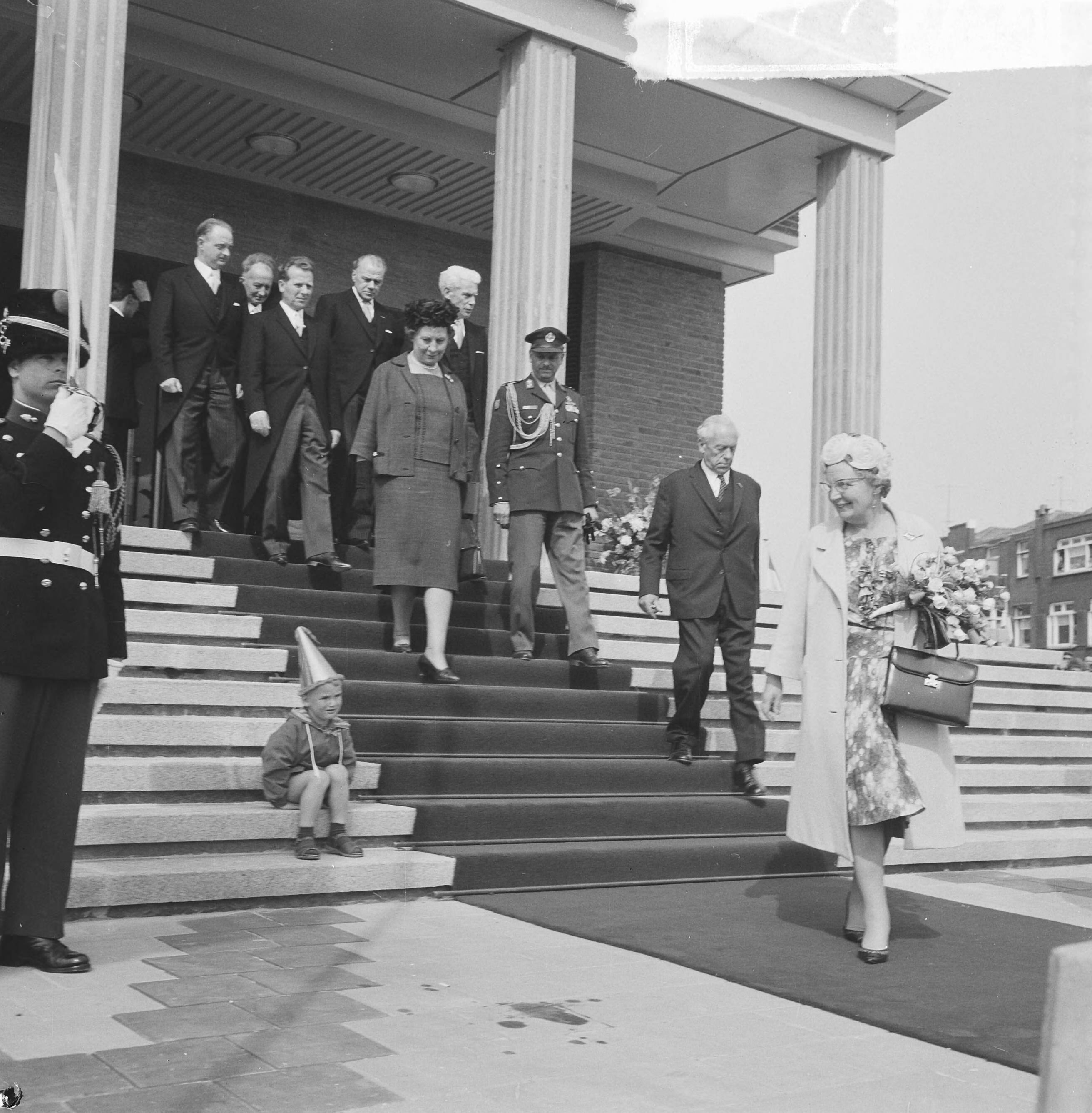 Koningin Juliana opent het nieuwe stadhuis te Vlissingen — Koningin Juliana is net de trappen van het stadhuis afgedaald. Links zit een jongetje met een feestmuts — Foto: Nijs, Jac. de / Anefo. Licentie: Set B: Rechtenvrij / Publiek Domein. Bron: Nationaal Archief. http://hdl.handle.net/10648/725899b3-1ebb-d266-7e85-c2ee8e9a2047