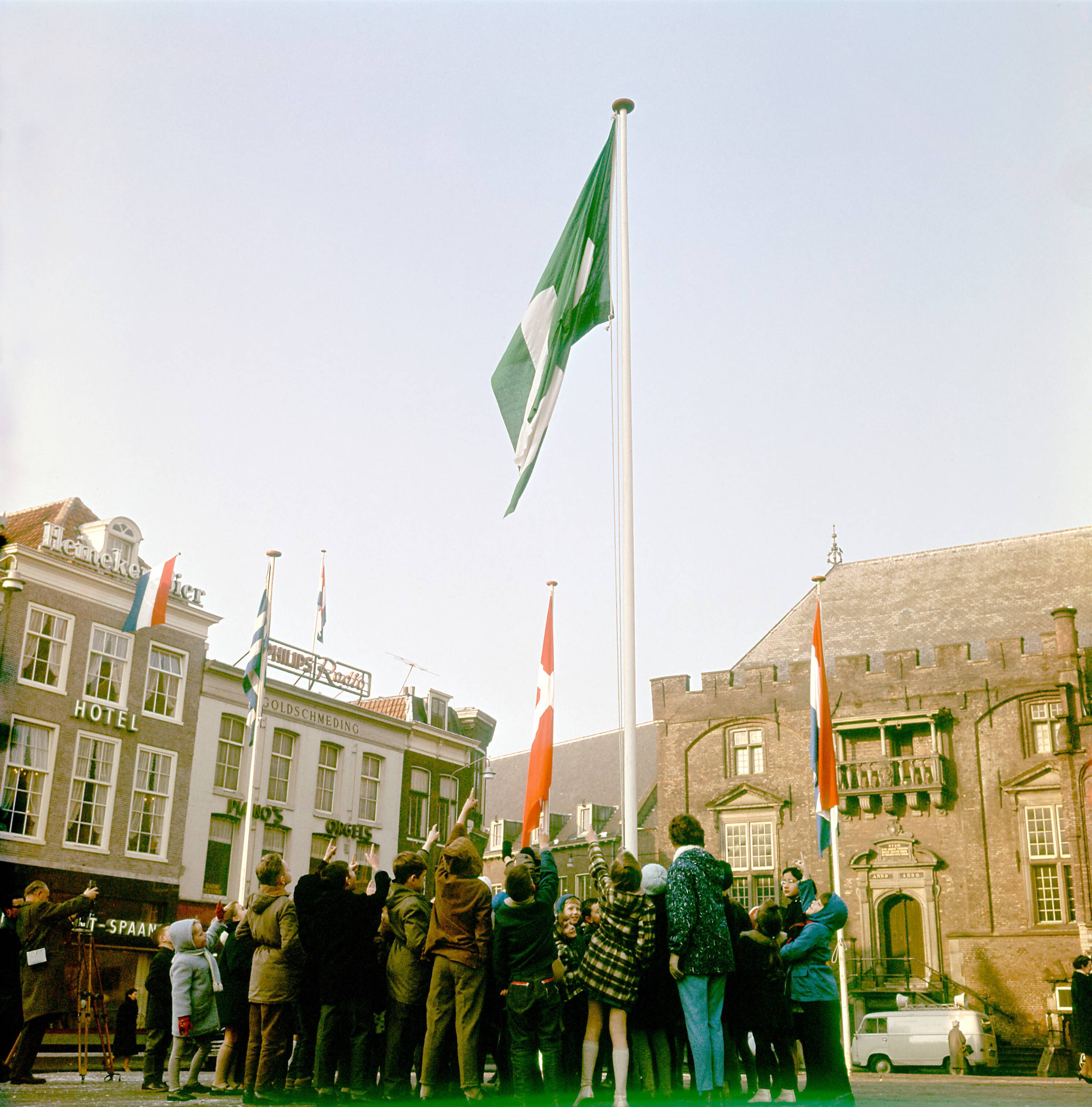 Collectie nader te bepalen — Op de Grote Markt te Haarlem wordt een groen-witte vlag gehesen, in aanwezigheid van kinderen. In de achtergrond het stadhuis. Links een hotel en een filiaal van de Piano- & Orgelhandel Goldschmeding — Foto: Fotograaf Onbekend / Fotobureau onbekend / Spaarnestad Photo (05887). Licentie: Set B: Rechtenvrij / Publiek Domein. Bron: Nationaal Archief. http://hdl.handle.net/10648/35429da7-a2ae-a382-e063-6df0900a6686