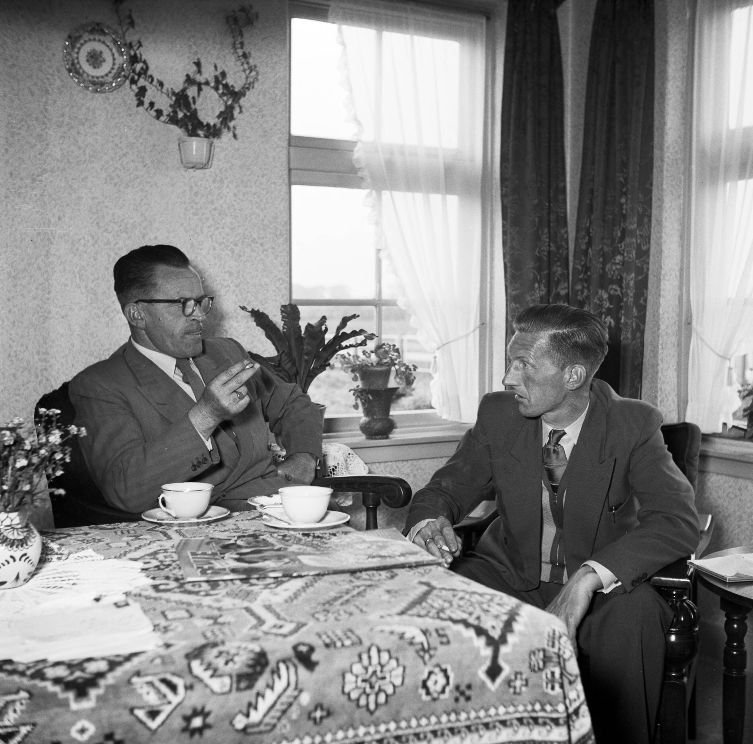 Fotocollectie Van de Poll — Gerard de Jong in gesprek met Ynse Postma, beide met een sigaret in de hand. Op tafel een exemplaar van het blad Revue — Foto: Poll, Willem van de. Licentie: Set B: Rechtenvrij / Publiek Domein. Bron: Nationaal Archief. http://hdl.handle.net/10648/aecce514-d0b4-102d-bcf8-003048976d84