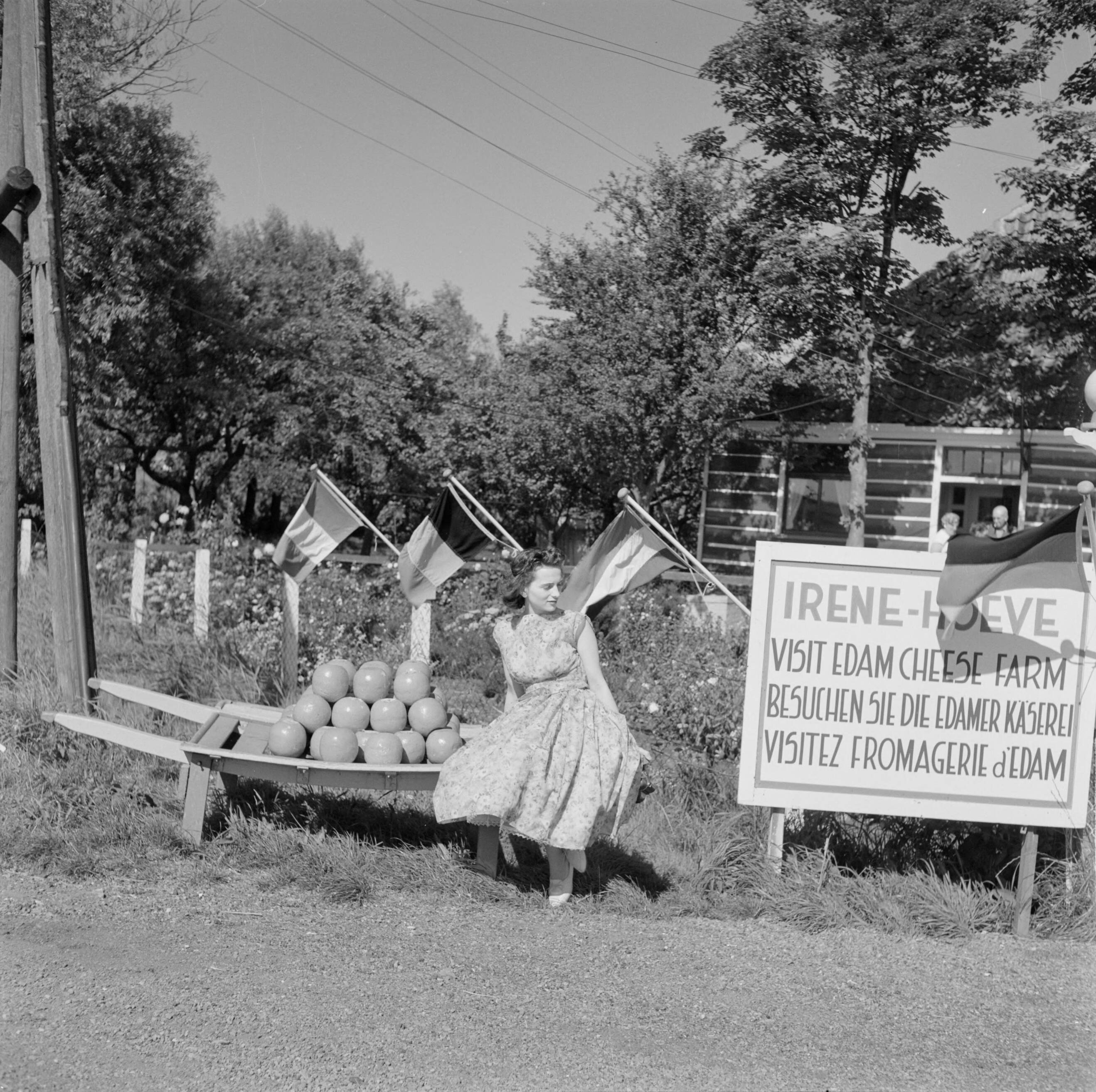 Volendam — Volendam. Irene-Hoeve: meisje zit op een berrie met kazen, naast een reclamebord in drie talen — Foto: Poll, Willem van de. Licentie: Set B: Rechtenvrij / Publiek Domein. Bron: Nationaal Archief. http://hdl.handle.net/10648/aecee1ca-d0b4-102d-bcf8-003048976d84