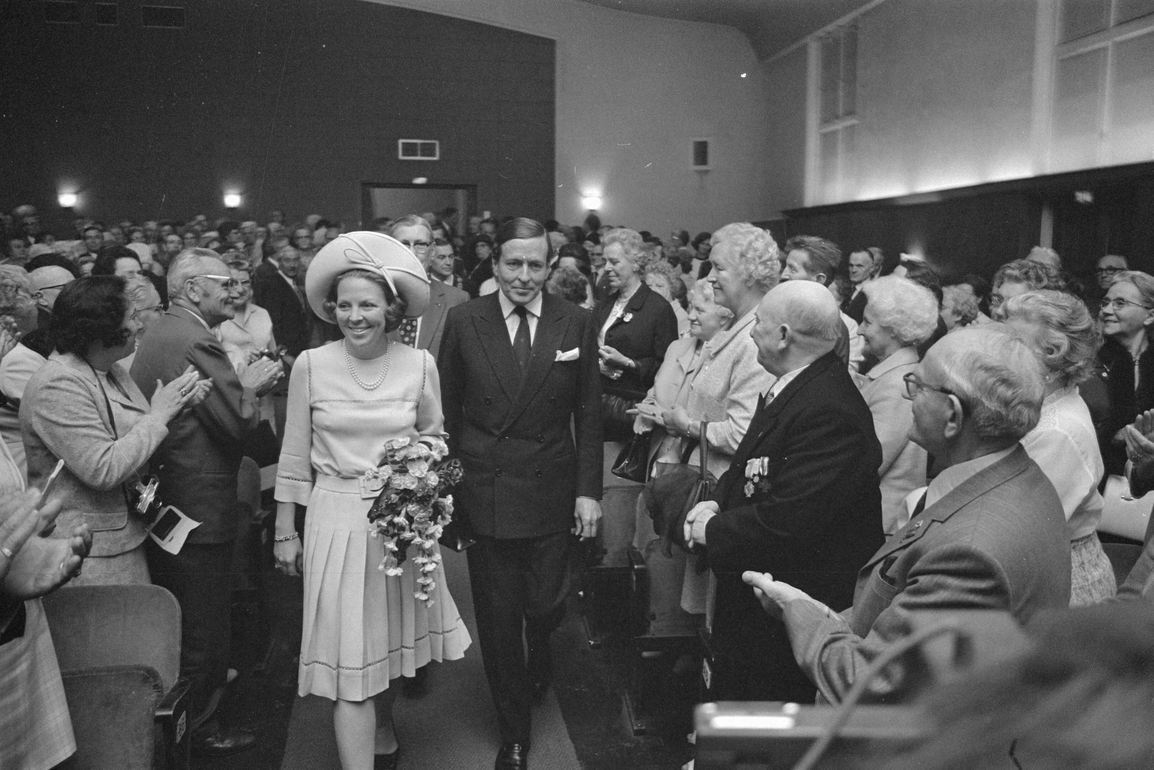 Aankomst van prinses Beatrix en prins Claus op het Expogé-congres in de KMA te Breda — Prinses Beatrix en prins Claus lopen onder applaus door de zaal — Foto: Fotograaf Onbekend / Anefo. Licentie: Set B: Rechtenvrij / Publiek Domein. Bron: Nationaal Archief. http://hdl.handle.net/10648/abc35678-d0b4-102d-bcf8-003048976d84