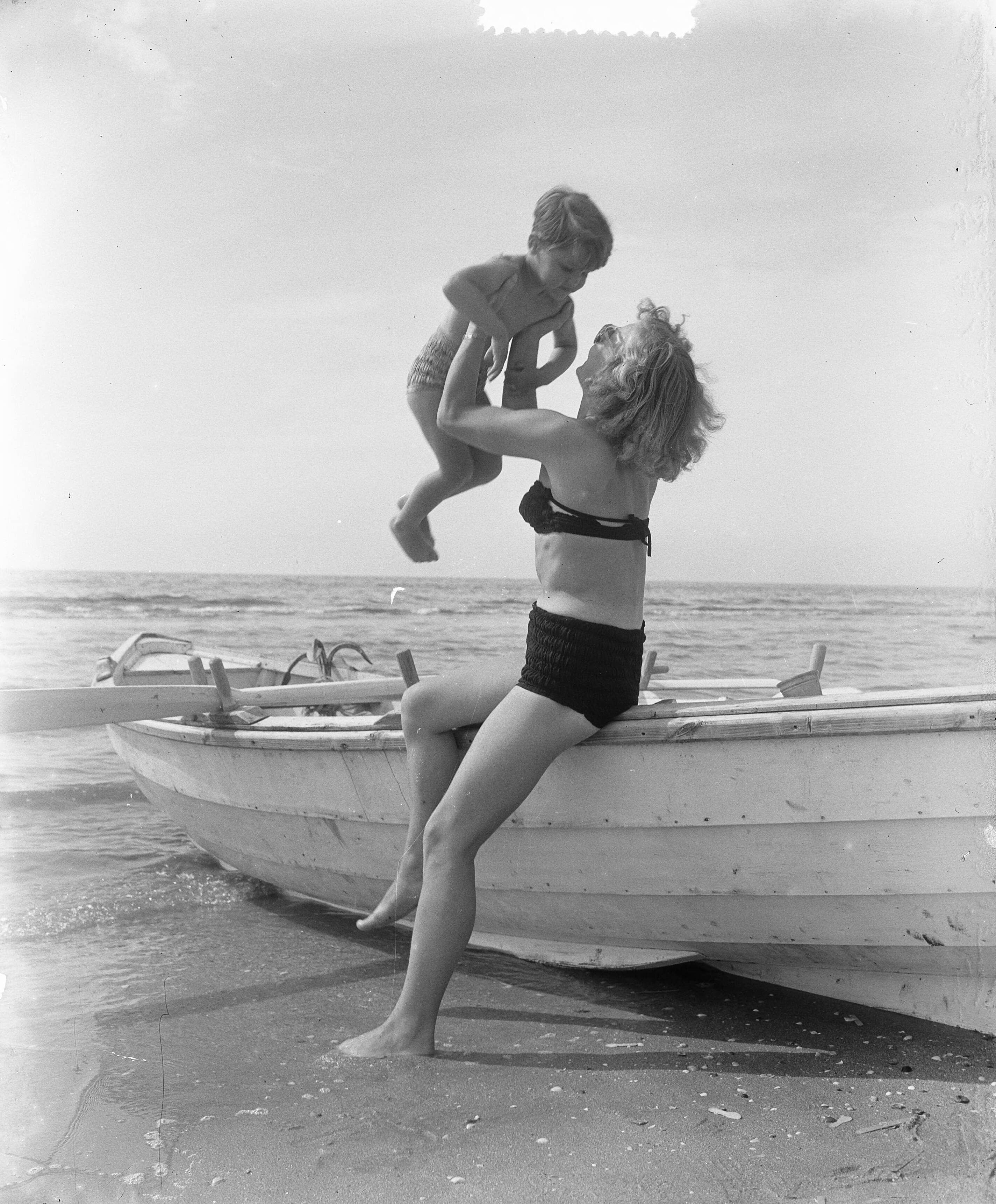 Fotocollectie Anefo — Zomerplaatjes aan strand van Zandvoort . Moeder tilt kind, zittend op een roeiboot — Foto: Pot, Harry / Anefo. Licentie: Set B: Rechtenvrij / Publiek Domein. Bron: Nationaal Archief. http://hdl.handle.net/10648/a90e564e-d0b4-102d-bcf8-003048976d84