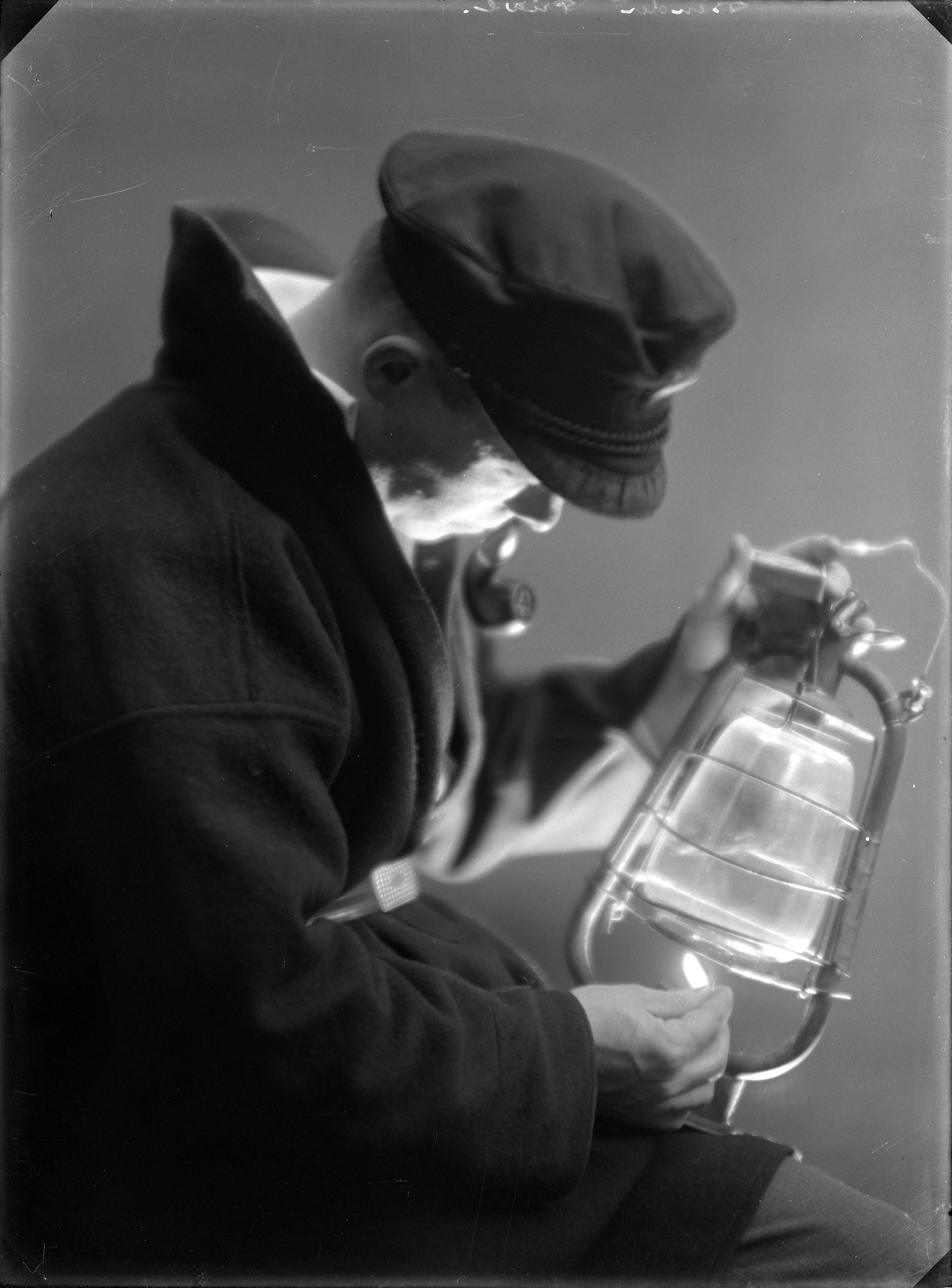 Portretten — Studio-opnames. Portret van een man in visserspak met pet, die een petroleum stormlamp aansteekt. Rechtsboven op glasnegatief: Bender privé. [Haarlem, Nederland,1918-1930]. — Foto: Berg, Th.C.J. van den / Spaarnestad Photo (05887). Licentie: Set B: Rechtenvrij / Publiek Domein. Bron: Nationaal Archief. http://hdl.handle.net/10648/35429da7-c548-a382-e063-6df0900a6686