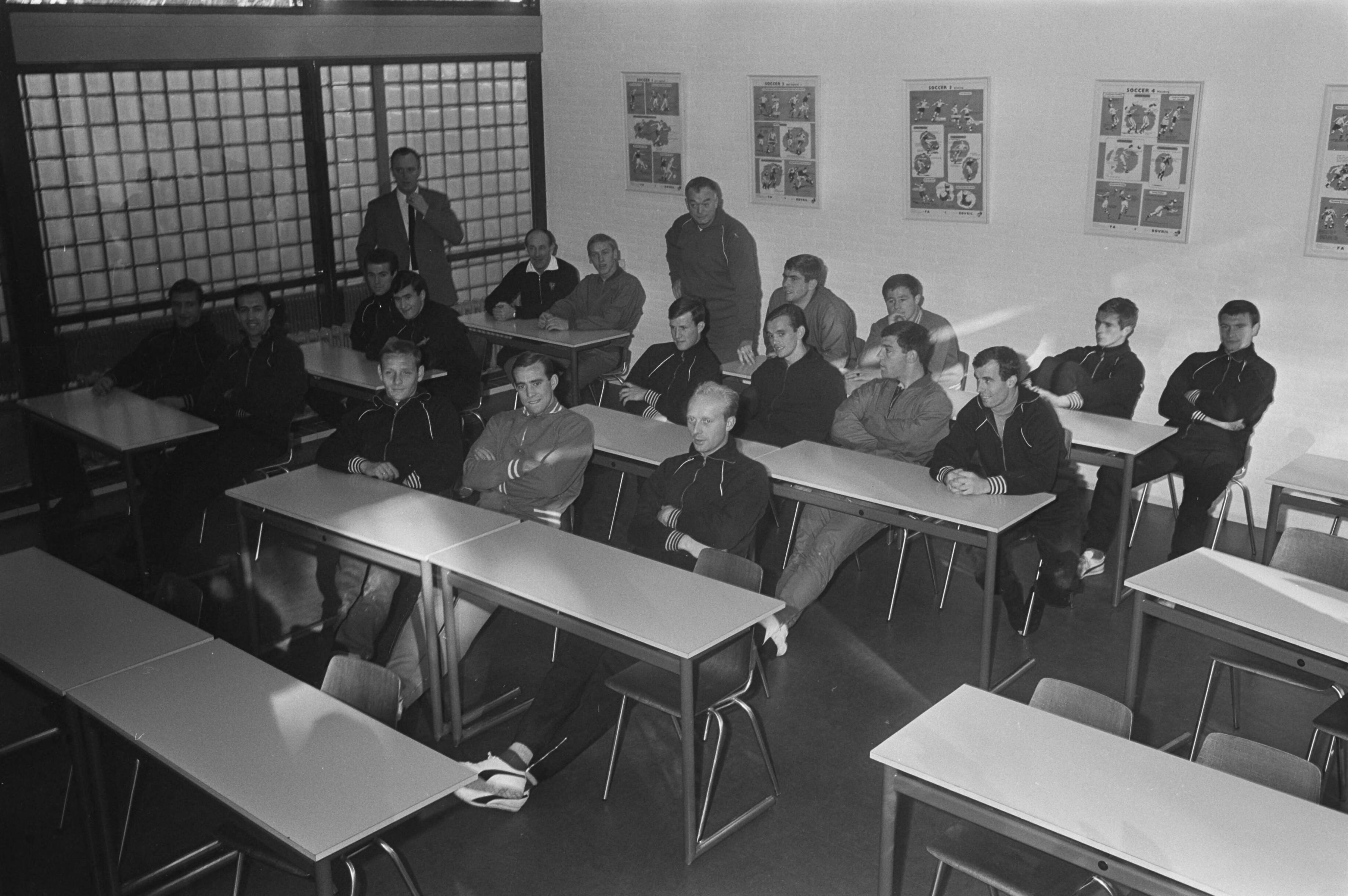 Training van het Nederlands Elftal — Training Nederlands Elftal in Zeist. Spelers tijdens de les in een klaslokaal. Staand achterin het klaslokaal: de trainer Denis Neville — Foto: Fotograaf Onbekend / Anefo. Licentie: Set B: Rechtenvrij / Publiek Domein. Bron: Nationaal Archief. http://hdl.handle.net/10648/aacbc2c8-d0b4-102d-bcf8-003048976d84