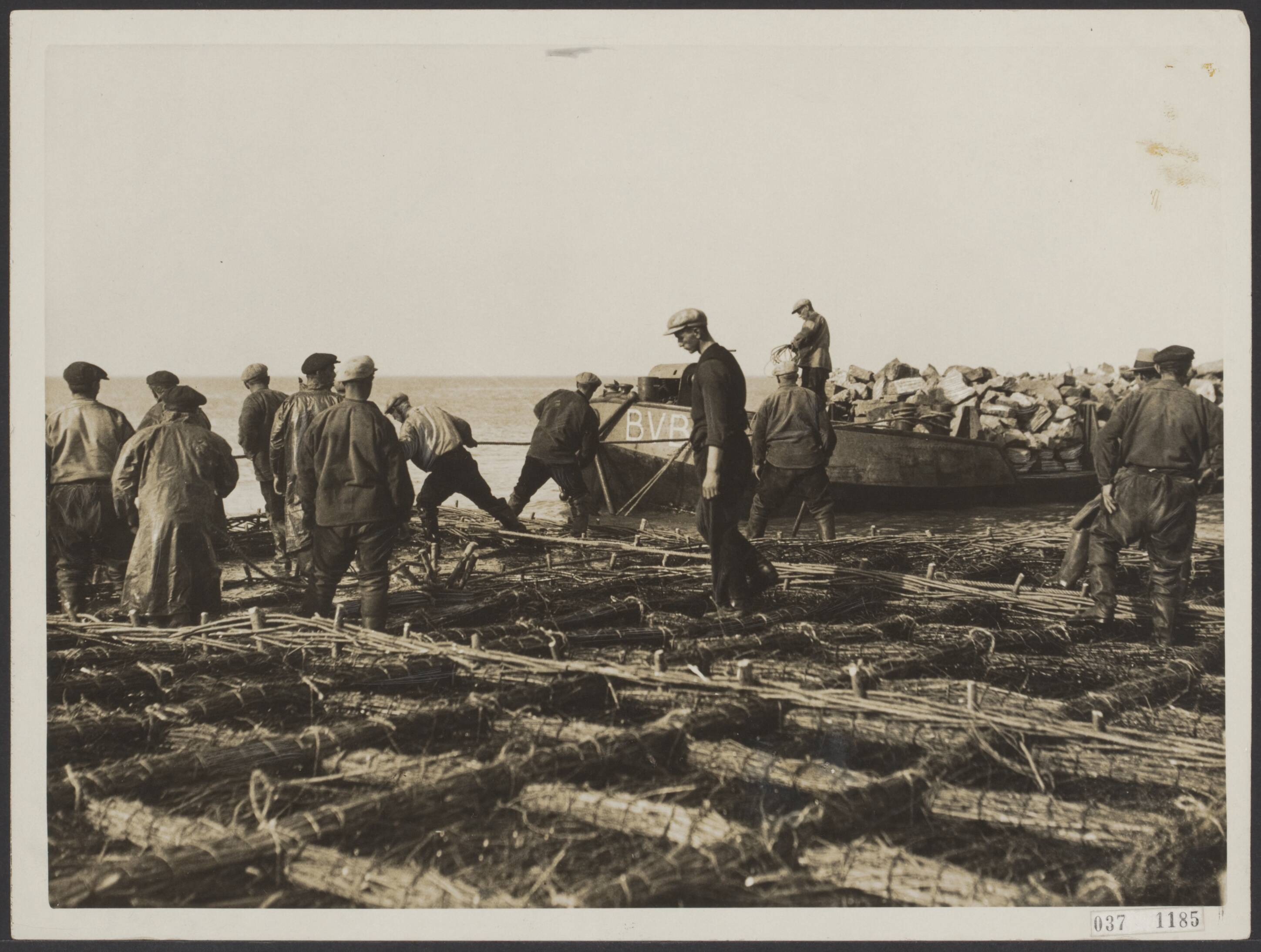 Fotocollectie Elsevier Binnenland — Een uit twijgen gemaakt zinkstuk wordt tot zinken gebracht hetwelk als fundering moet dienen voor de te Scheveningen te bouwen nieuwe golfbreker. Een schuit met bazaltklinkers, voor heet verzwaren, wordt langszij getrokken — Foto: N.V. Polygoon. Licentie: Set B: Rechtenvrij / Publiek Domein. Bron: Nationaal Archief. http://hdl.handle.net/10648/adefcab2-d0b4-102d-bcf8-003048976d84