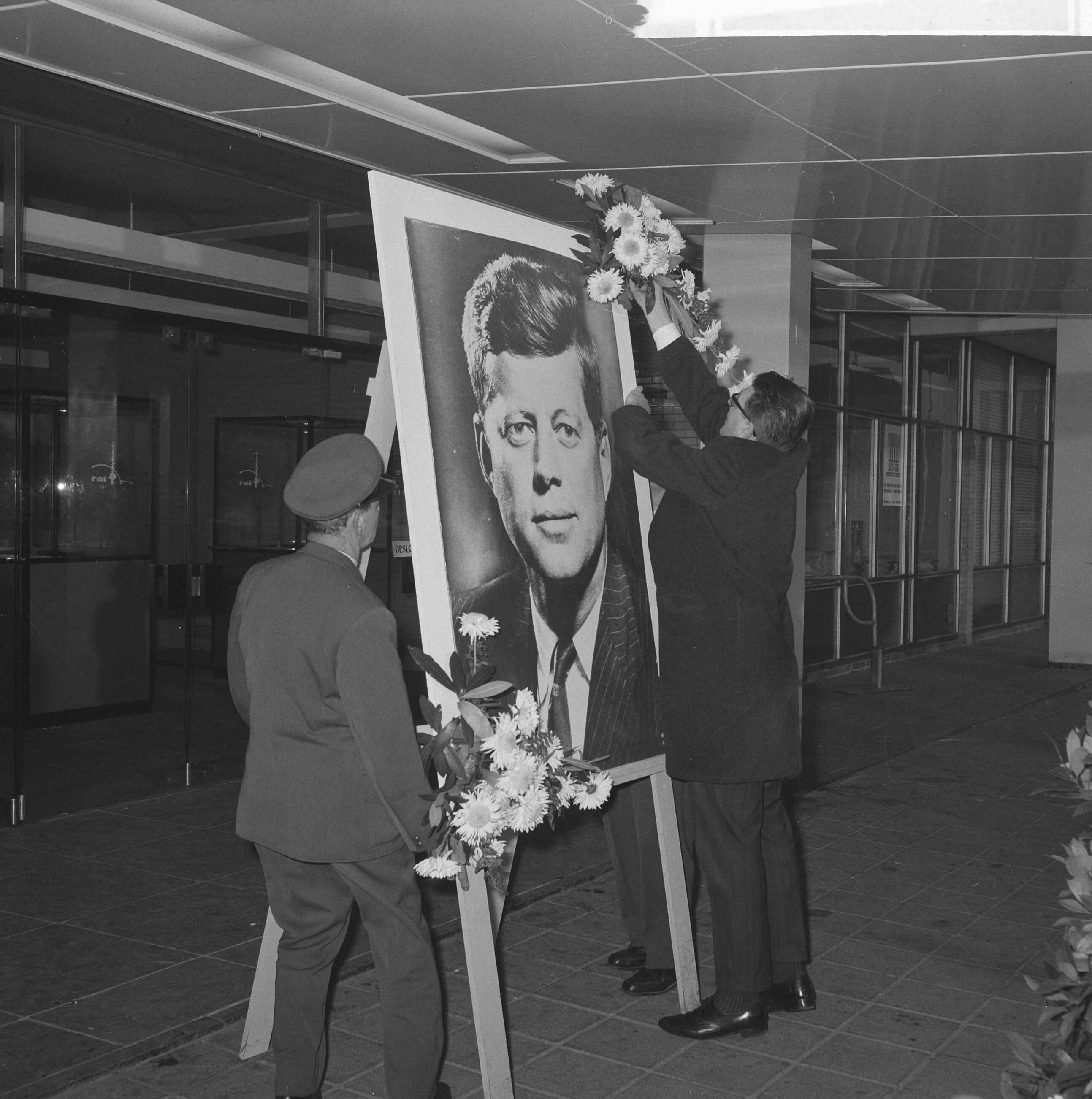 Fotocollectie Anefo — Herdenkingsplaats voor president Kennedy bij het RAI-gebouw te Amsterdam. Bloemen worden aan zijn portret bevestigd — Foto: Nijs, Jac. de / Anefo. Licentie: Set B: Rechtenvrij / Publiek Domein. Bron: Nationaal Archief. http://hdl.handle.net/10648/aa4f6ac0-d0b4-102d-bcf8-003048976d84