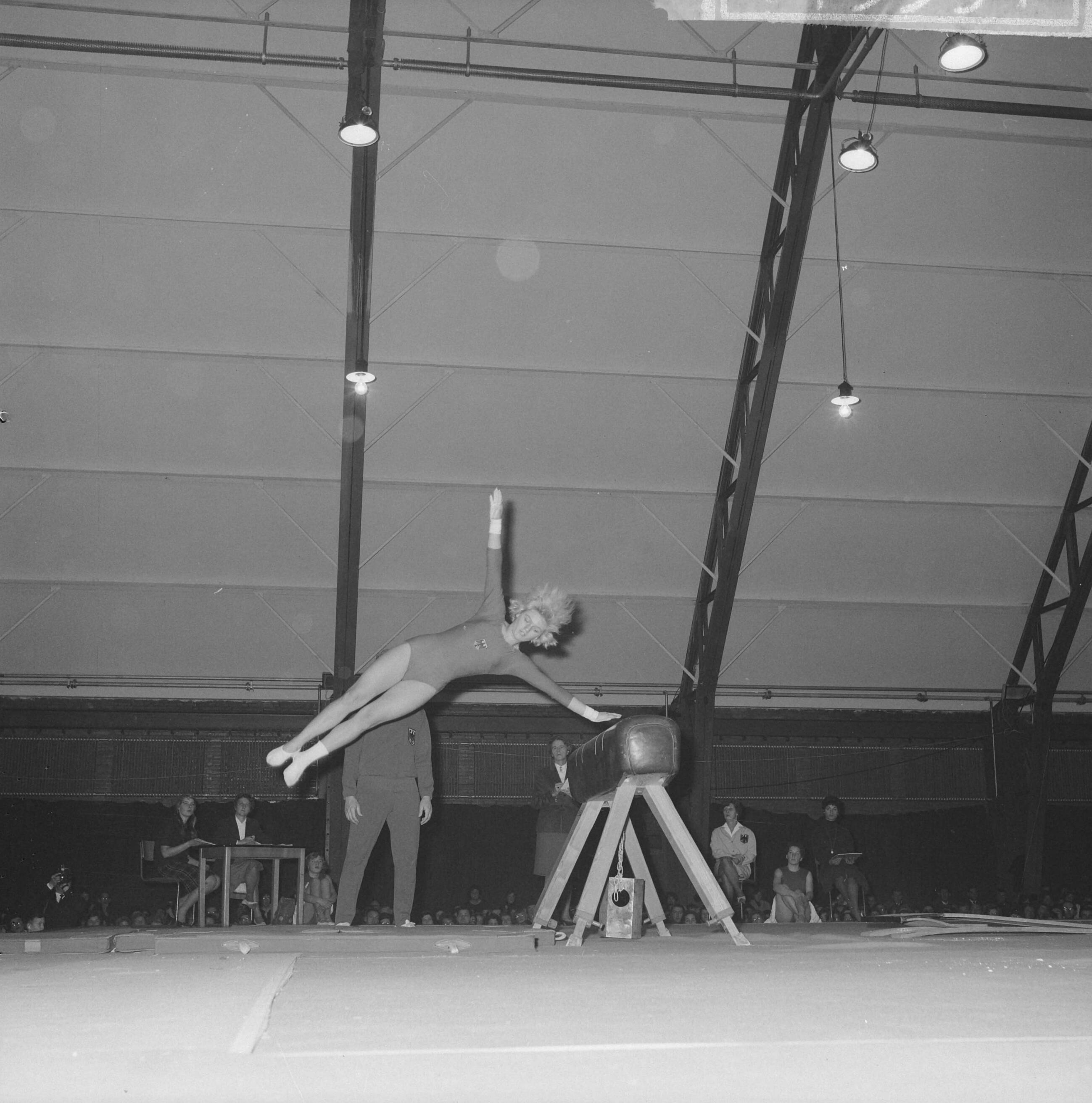 Fotocollectie Anefo — Turninterland Nederland-West-Duitsland in de Centrale Veilinghal te Aalsmeer. Een Duitse turnster bij paardsprong. In de achtergrond trainer Niko Szabo — Foto: Nijs, Jac. de / Anefo. Licentie: Set B: Rechtenvrij / Publiek Domein. Bron: Nationaal Archief. http://hdl.handle.net/10648/aa200438-d0b4-102d-bcf8-003048976d84