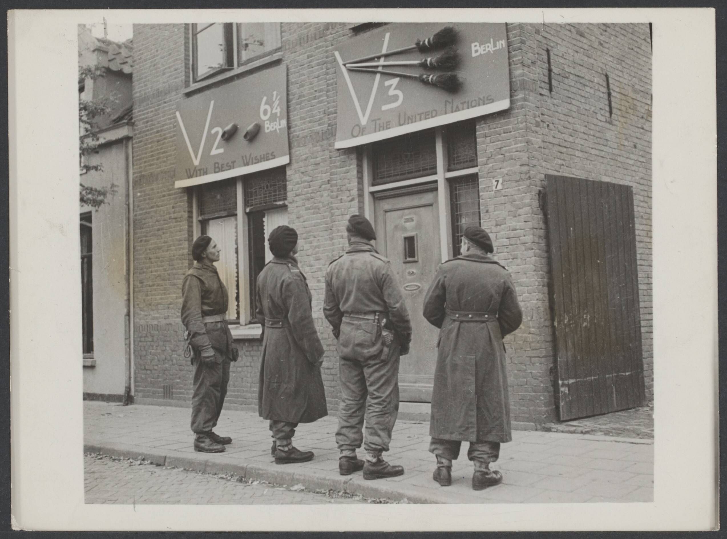 Bevrijding Nederland 1944 [bevat ook foto's uit 1945] — Breda. Vier Poolse militairen kijken naar twee platen bevestigd aan een woonhuis. Het linker heeft het opschrift "V2 6 1/4 Berlin With Best Wishes", met twee vastgemaakte klompen. Het rechter "V3 Berlin. Of the United Nations". Met drie vastgemaakte bezems — Foto: Keystone Press / Rechtenvrij. Licentie: Set B: Rechtenvrij / Publiek Domein. Bron: Nationaal Archief. http://hdl.handle.net/10648/11c8ed31-5c5d-7cd1-6f4f-327e2e8f831a