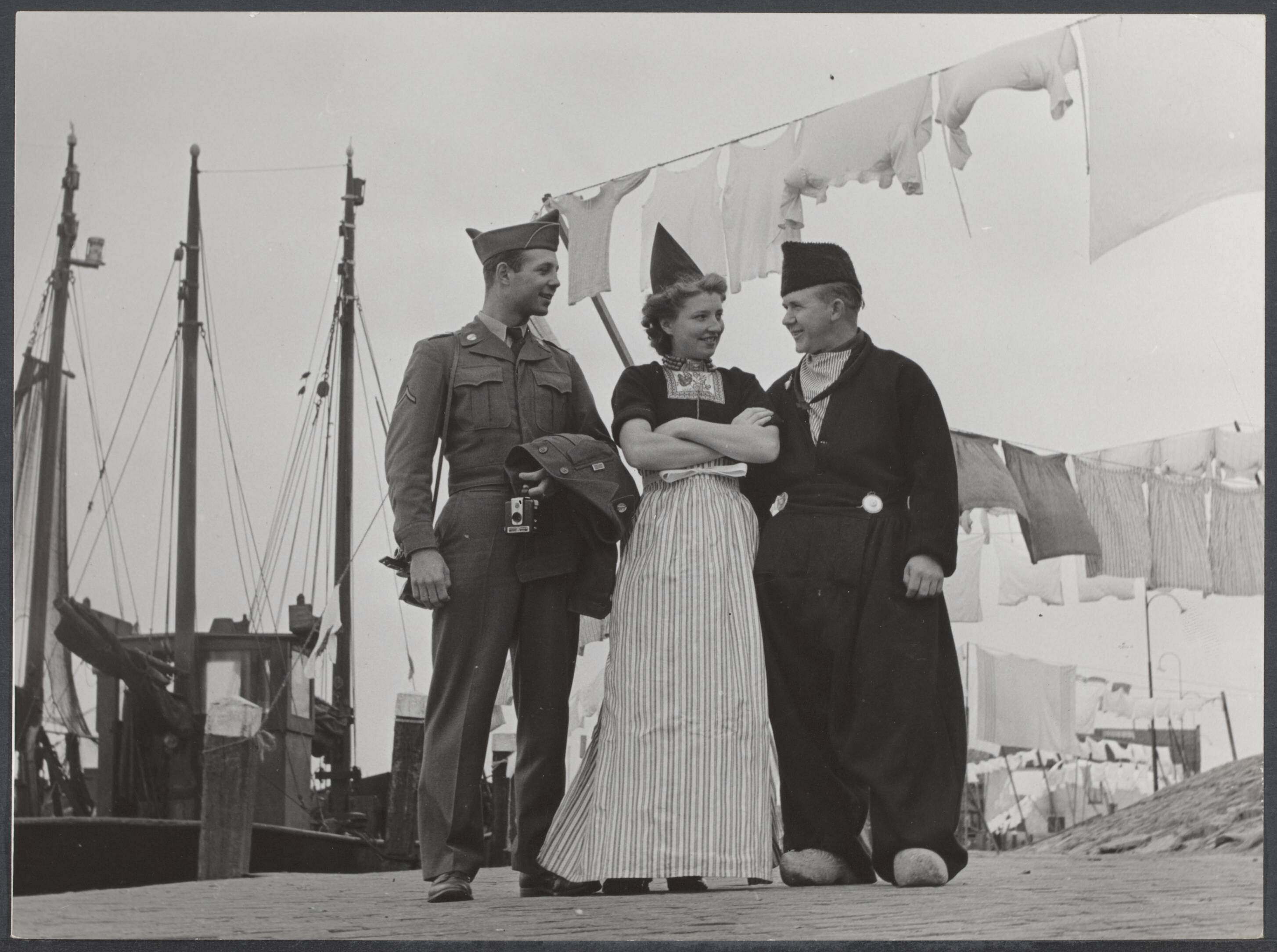 Soldatenleven buiten dienst - Nederland — Geen bijschrift [Amerikaanse militair met fototoestel poseert in een haven (Volendam) met een man en vrouw in klederdracht, achter hen visserschapen en wasgoed aan waslijnen] — Foto: Pers Foto Dienst / Rechtenvrij. Licentie: Set B: Rechtenvrij / Publiek Domein. Bron: Nationaal Archief. http://hdl.handle.net/10648/5525148c-5b4c-056c-1076-9aead811e3e3
