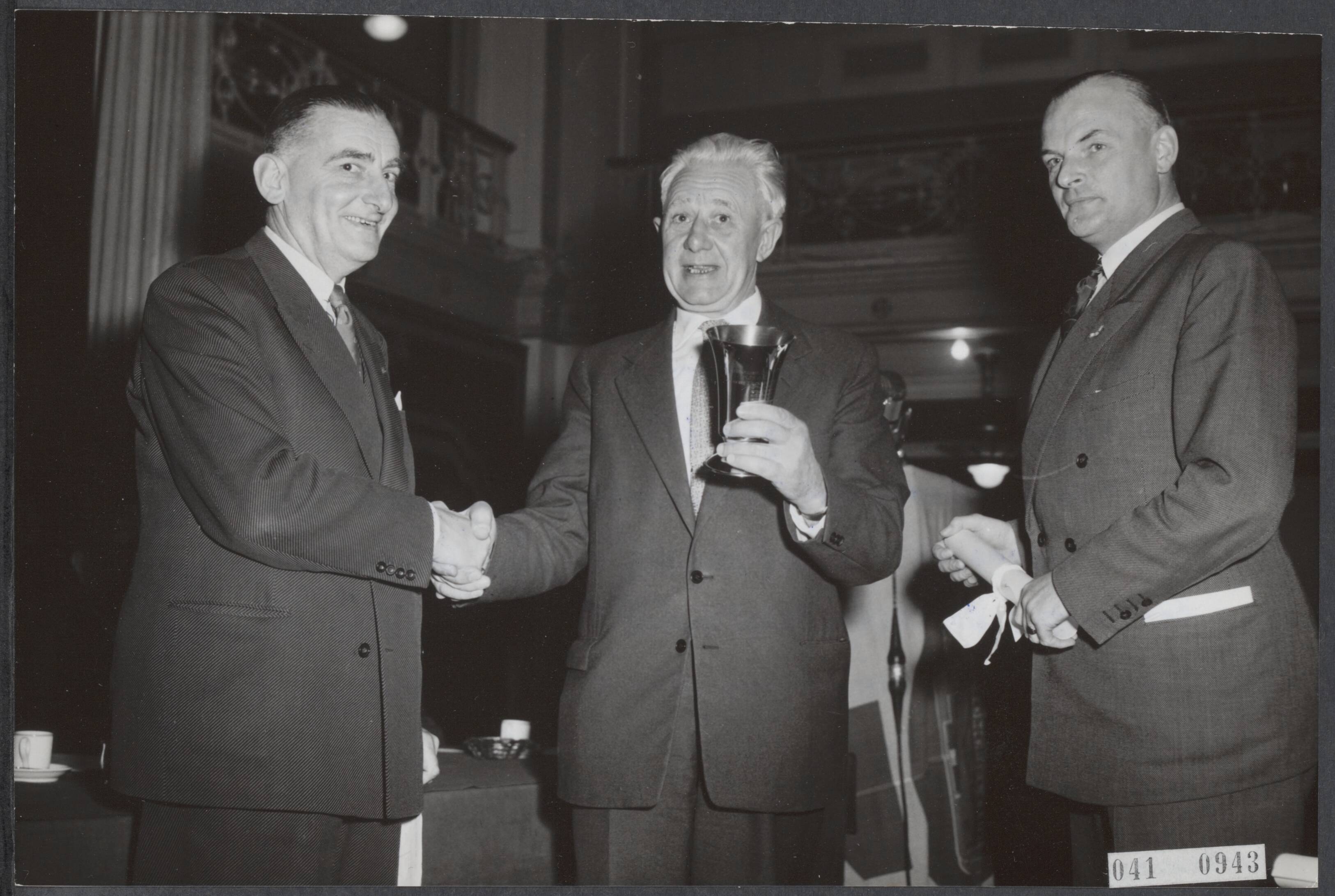 Fotocollectie Elsevier Binnenland — De minister van Waterstaat mr. J. Algera heeft in het Kurhaus te Scheveningen de jaarlijkse Wegverkeersdag geopend. De procureur-generaal van het Gerechtshof te Den Haag, mr. D.J. van Gilsen (liks) reikte aan de hoofdcommissaris van Amsterdam (rechts) een oorkonde uit, daar Amsterdam in 1955 de minste verkeersongevallen noteerde van de drie grote gemeenten. In het midden de loco-burgemeester van Den Haag C.H.P.W. van den Oever die de beker in ontvangst nam, daar Den Haag in zes jaar tijd procentsgewijs de minste ongevallen noteerde — Foto: Fotograaf Onbekend / Anefo. Licentie: Set B: Rechtenvrij / Publiek Domein. Bron: Nationaal Archief. http://hdl.handle.net/10648/adf735c2-d0b4-102d-bcf8-003048976d84