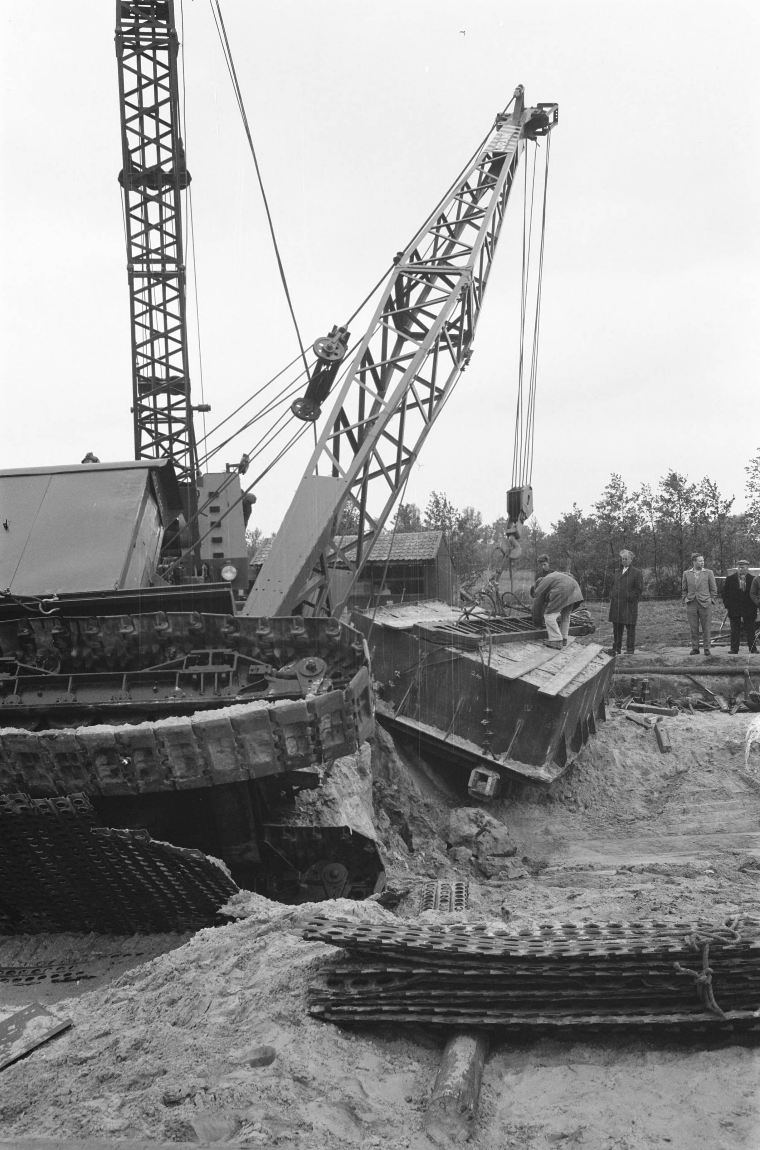 Fotocollectie Anefo — In Esch (N.B.) zijn militairen van het depot Genietroepen van de Koning Willem I-kazerne in Den Bosch bezig geweest met het uitgraven van een Romeins graf uit het begin van onze jaartelling. Het graf werd in een metalen bekisting gebouwd om in zijn geheel naar Amersfoort te worden overgebracht, waar de Rijksdienst voor Oudheidkundig Bodemonderzoek het zal bestuderen. Gisteren wilden twee kranen het graf op een vrachtauto tillen; een van de kranen kantelde echter. Het graf bleef ongedeerd — Foto: Pot, Harry / Anefo. Licentie: Set B: Rechtenvrij / Publiek Domein. Bron: Nationaal Archief. http://hdl.handle.net/10648/a9d66562-d0b4-102d-bcf8-003048976d84