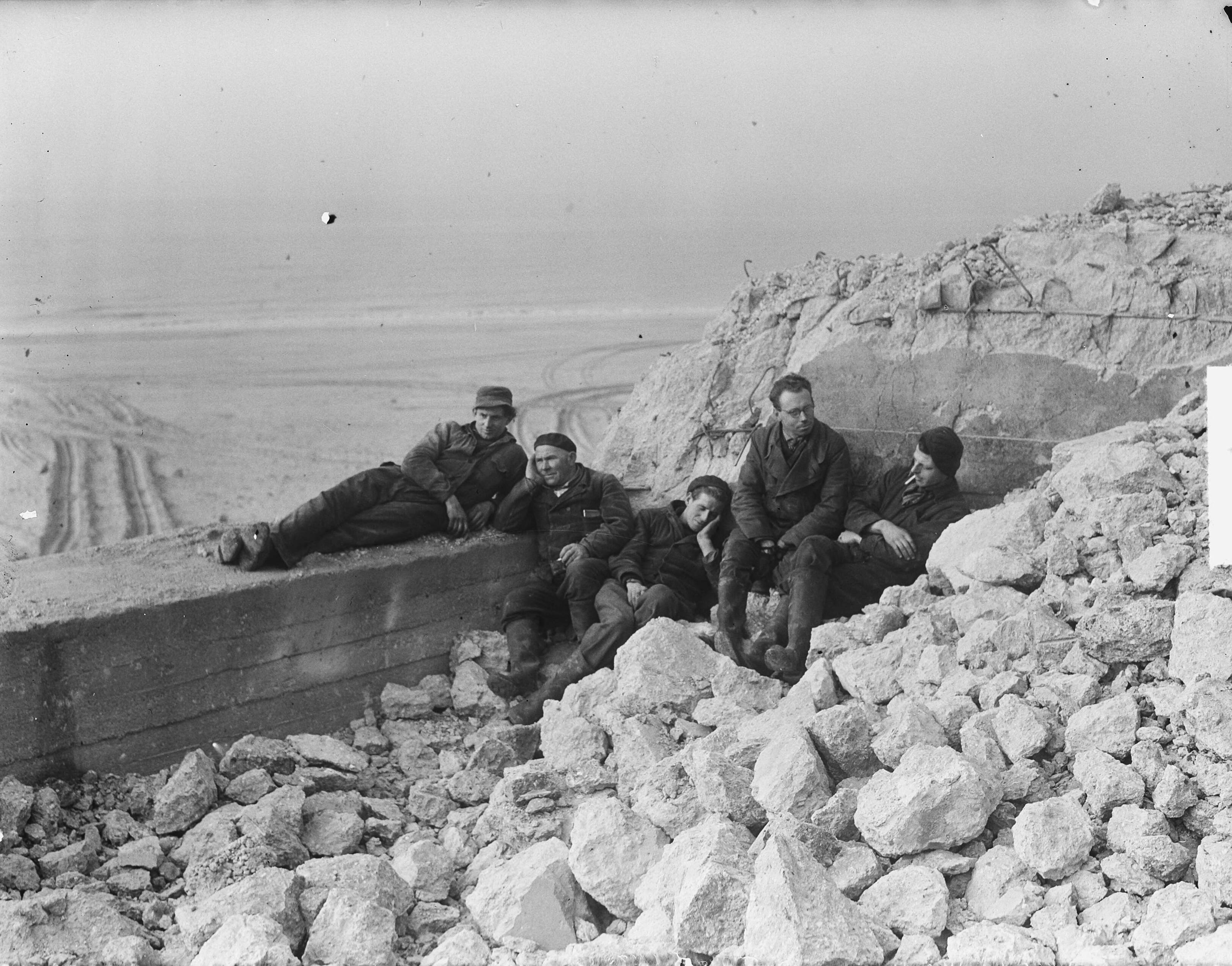 Arbeiders pauzeren in de zon tijdens de sloop van bunkers te Zandvoort — Foto: Vogel, Carel L. de / Anefo. — Licentie: Set B: Rechtenvrij / Publiek Domein. Bron: Nationaal Archief. http://hdl.handle.net/10648/a8e5bcac-d0b4-102d-bcf8-003048976d84