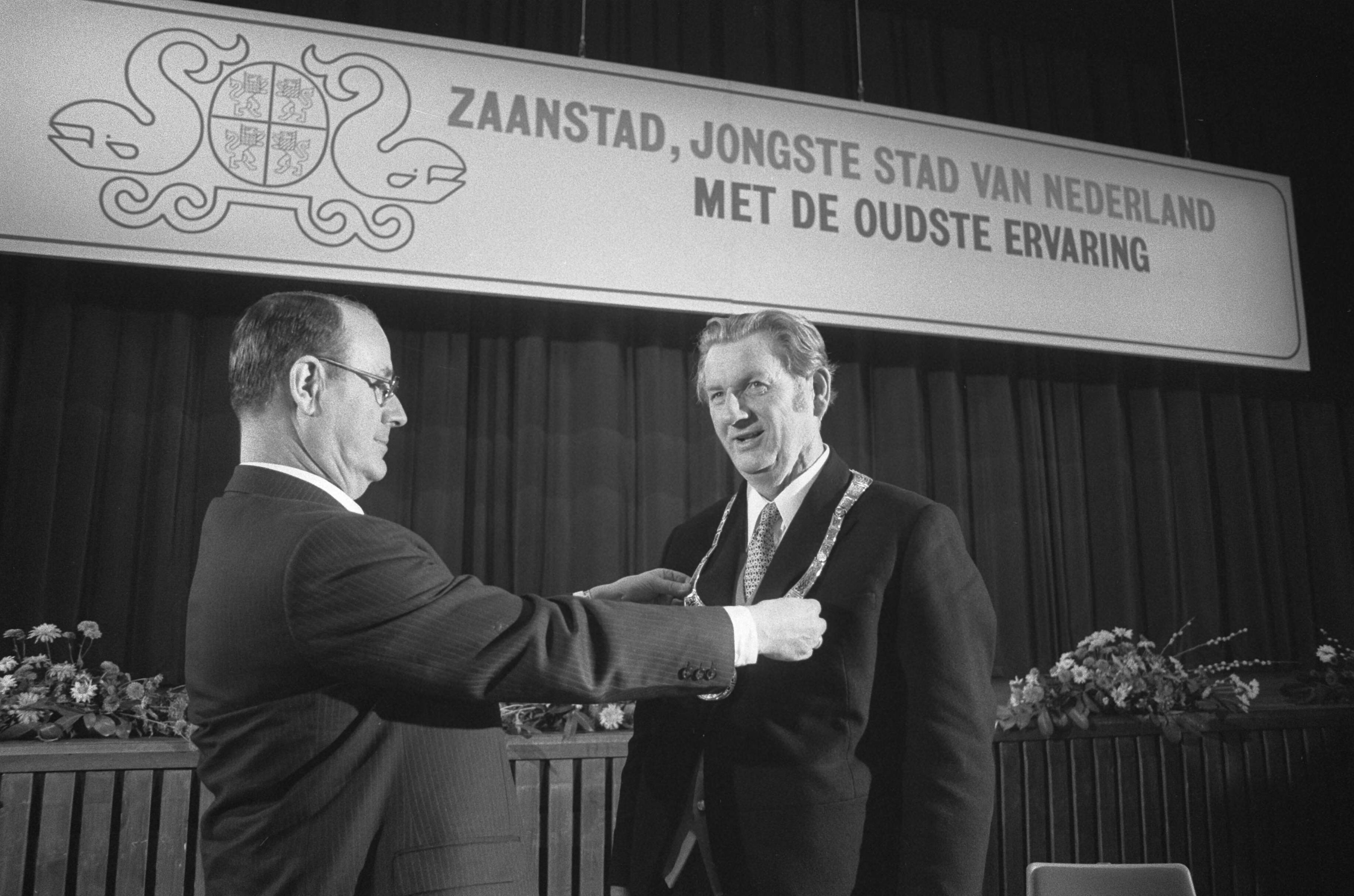 Burgemeester R. Laan (rechts) wordt geïnstalleerd als burgemeester van Zaanstad — Mr. Kranenburg (Commissaris der Koningin) hangt de nieuw geïnstalleerde burgemeester de ambtsketen om — Foto: Fotograaf Onbekend / Anefo. Licentie: Set B: Rechtenvrij / Publiek Domein. Bron: Nationaal Archief. http://hdl.handle.net/10648/ac3fd036-d0b4-102d-bcf8-003048976d84