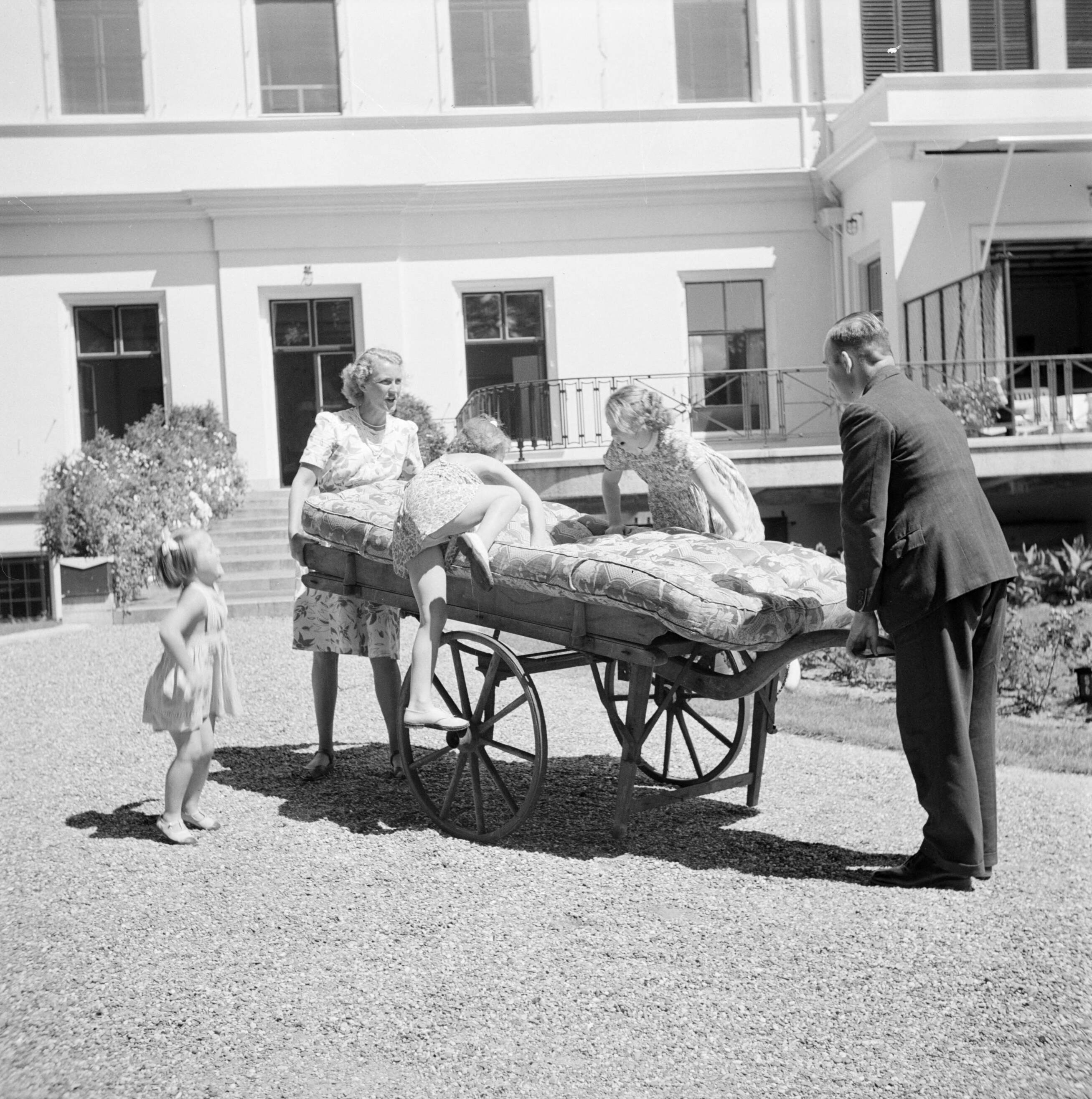 De koninklijke familie op paleis Soestdijk — De prinsessen Beatrix en Irene klimmen voor de achtergevel van het paleis op een handkar die door personeel wordt vastgehouden. Prinses Margriet kijkt toe. — Foto: Poll, Willem van de. Licentie: Set B: Rechtenvrij / Publiek Domein. Bron: Nationaal Archief. http://hdl.handle.net/10648/aee98b38-d0b4-102d-bcf8-003048976d84