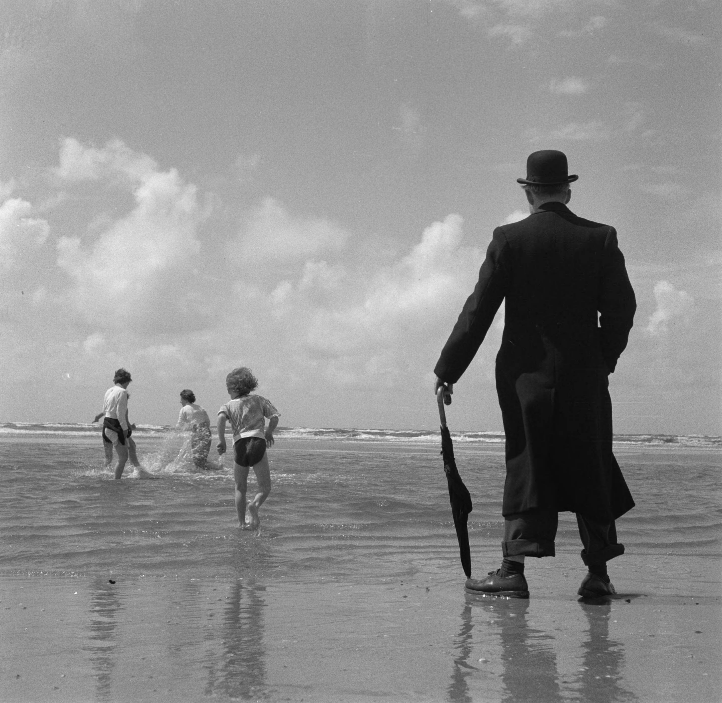 Fotocollectie Anefo — Zandvoort juni 1956. Een geklede oudere heer met paraplu en bolhoed kijkt naar spelende kinderen op het strand — Foto: Pot, Harry / Anefo. Licentie: Set B: Rechtenvrij / Publiek Domein. Bron: Nationaal Archief. http://hdl.handle.net/10648/a94a379a-d0b4-102d-bcf8-003048976d84