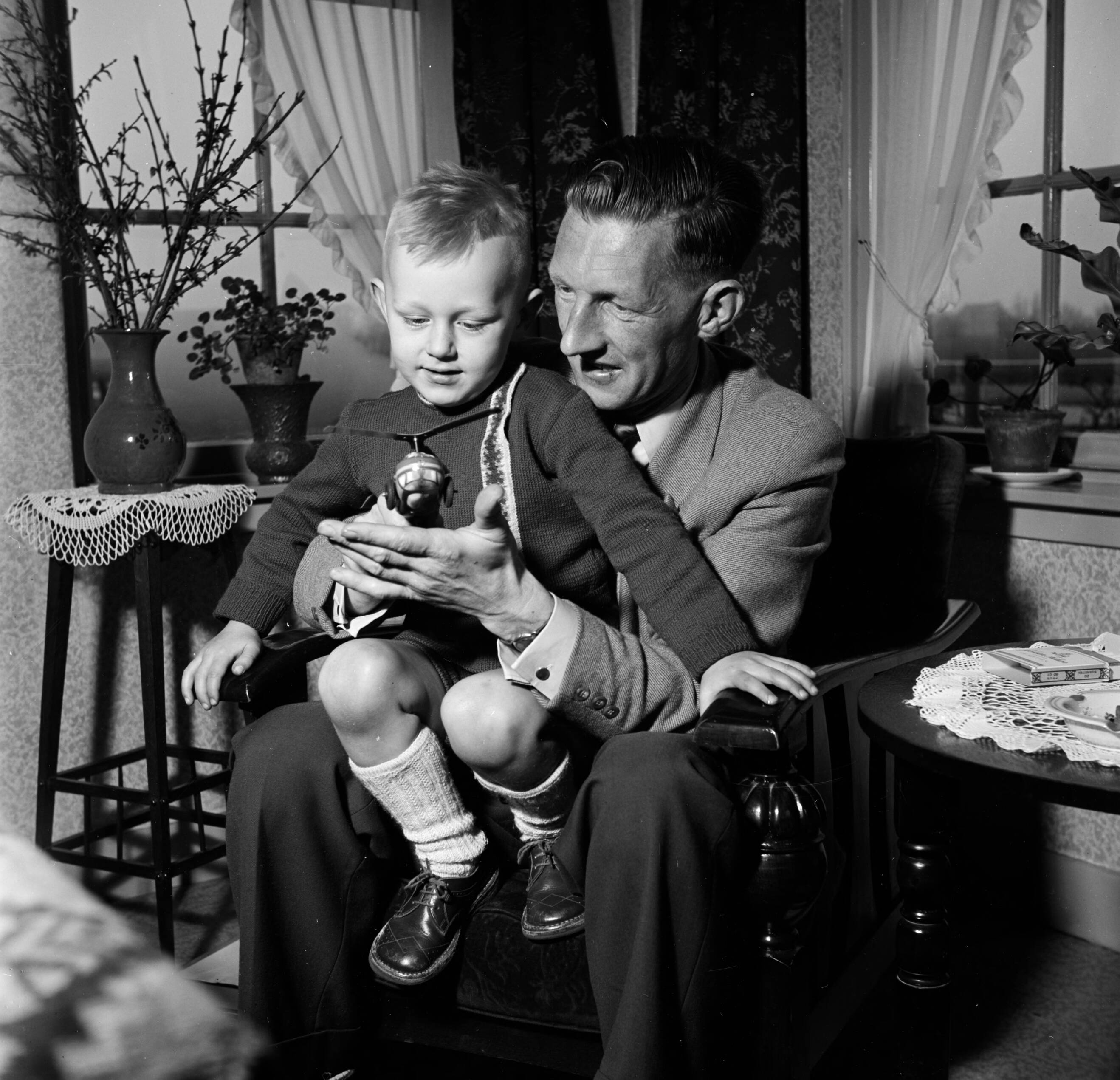 Fotocollectie Van de Poll — Gerard met zijn vierjarig zoontje Gerrie op schoot met een speelgoed helikopter. Links en bloementafeltje met een vaas met uitbottende takken — Foto: Poll, Willem van de. Licentie: Set B: Rechtenvrij / Publiek Domein. Bron: Nationaal Archief. http://hdl.handle.net/10648/aecccffc-d0b4-102d-bcf8-003048976d84