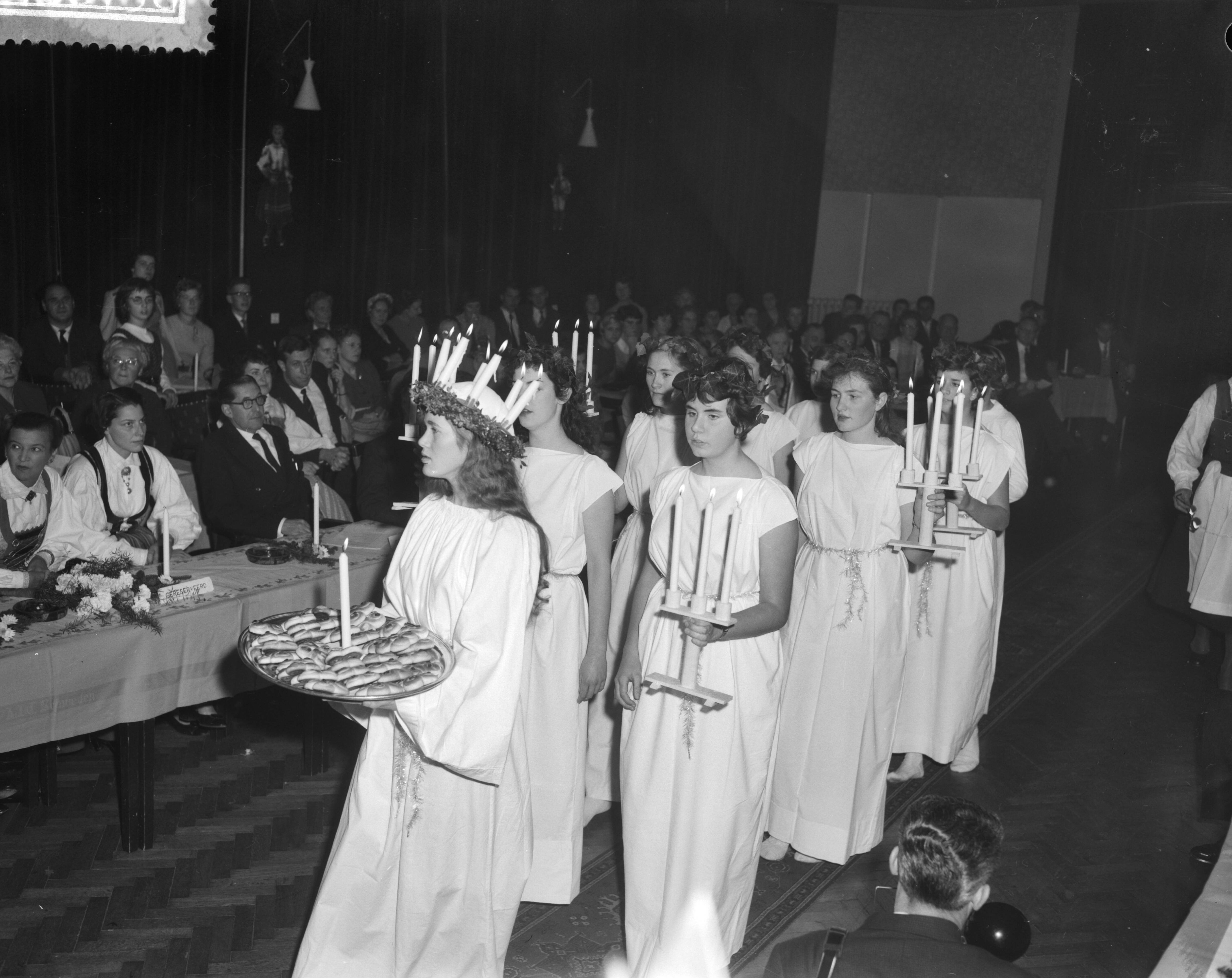 Fotocollectie Anefo — Viering van het Luciafeest door de Zweeds Nederlandse Vereniging in de Dierentuin te Den Haag, Lucia (de 18-jarige Helbertijn Küller uit Den Haag) gevolgd door "ternin" (dienstmaageden) komt de zaal binnen — Foto: Behrens, Herbert F. / Anefo. Licentie: Set B: Rechtenvrij / Publiek Domein. Bron: Nationaal Archief. http://hdl.handle.net/10648/a9a3abb8-d0b4-102d-bcf8-003048976d84