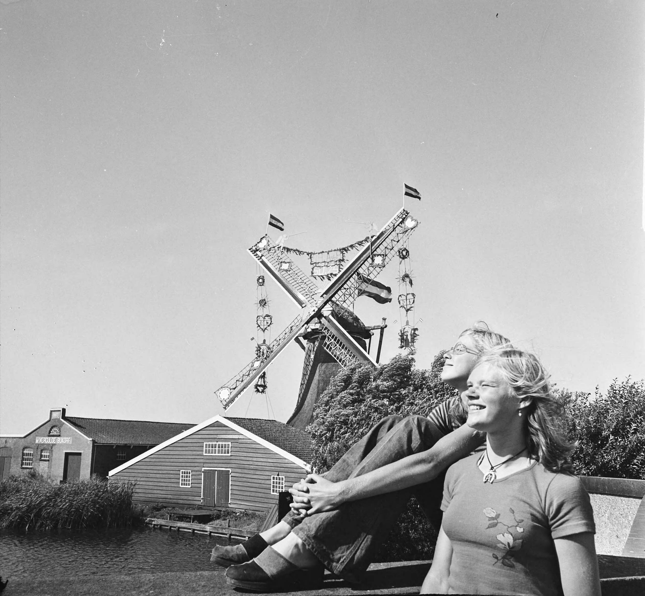 Molen in bruidstooi in Westzaan i.v.m. een tentoonstelling — Twee jongeren met op de achtergrond de met vlaggen en slingers versierde molen — Foto: Mieremet, Rob / Anefo. Licentie: Set B: Rechtenvrij / Publiek Domein. Bron: Nationaal Archief. http://hdl.handle.net/10648/ac6eea88-d0b4-102d-bcf8-003048976d84