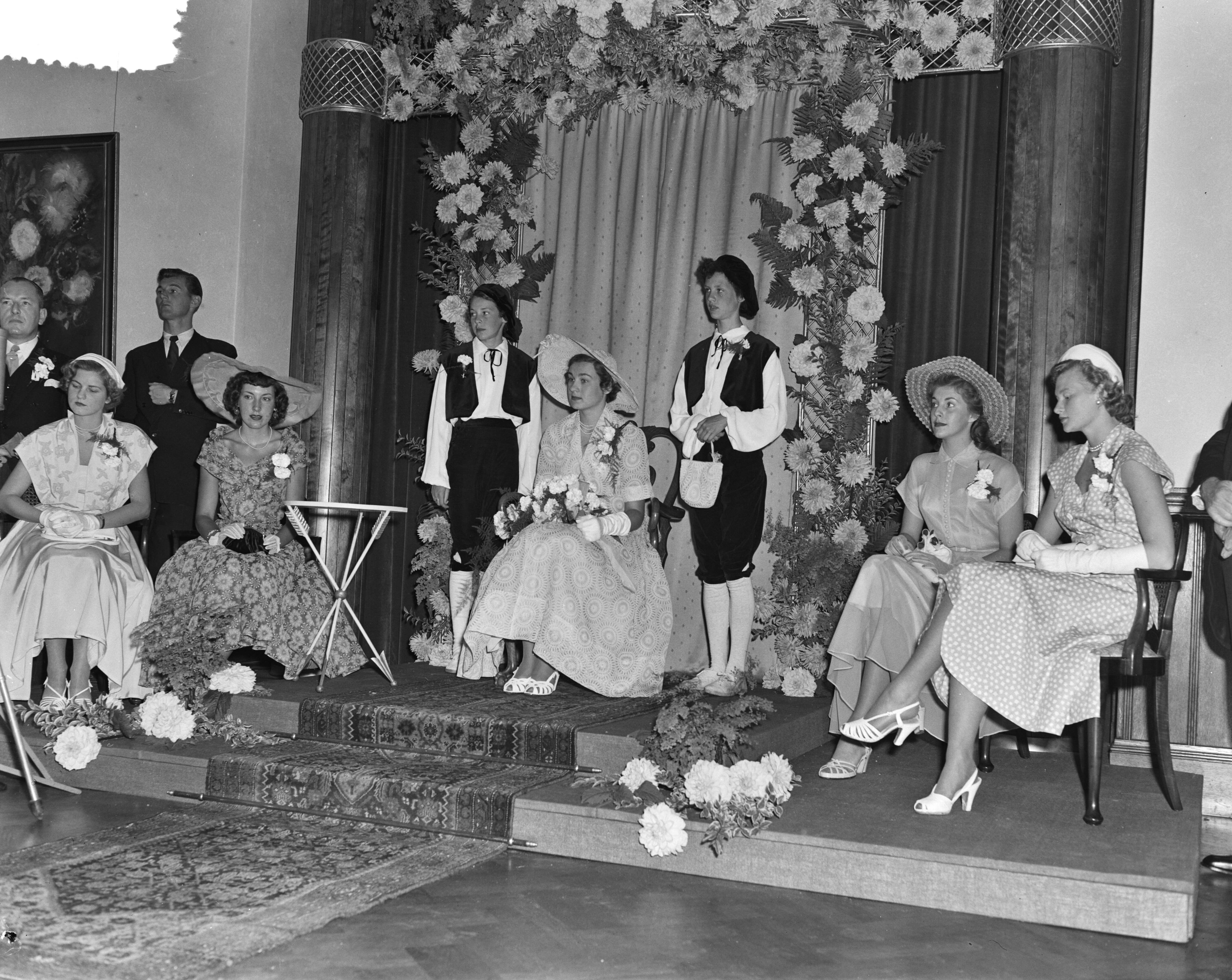 Intocht Bloemenkoningin van Den Haag — Intocht Bloemenkoningin van Den Haag. Zij zit in de oude raadszaal geflankeerd door pages en hofdames — Foto: Pot, Harry / Anefo. Licentie: Set B: Rechtenvrij / Publiek Domein. Bron: Nationaal Archief. http://hdl.handle.net/10648/a8edcbae-d0b4-102d-bcf8-003048976d84