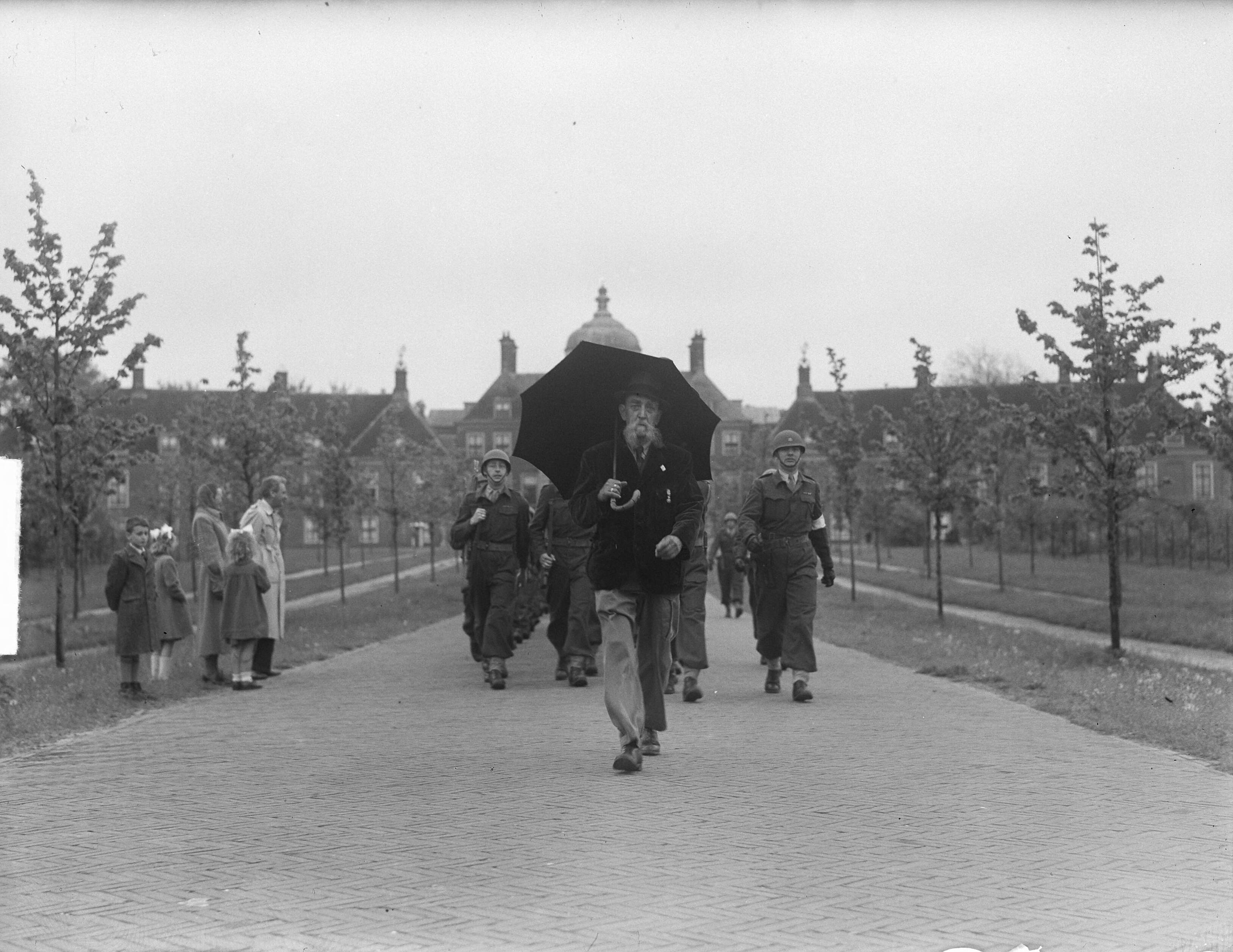NVJ mars te Scheveningen — NJV mars te Scheveningen [oudere wandelaar met paraplu voor paleis Huis ten Bosch met achter zich een groep militairen] — Foto: Pot, Harry / Anefo. Licentie: Set B: Rechtenvrij / Publiek Domein. Bron: Nationaal Archief. http://hdl.handle.net/10648/a8d8b872-d0b4-102d-bcf8-003048976d84