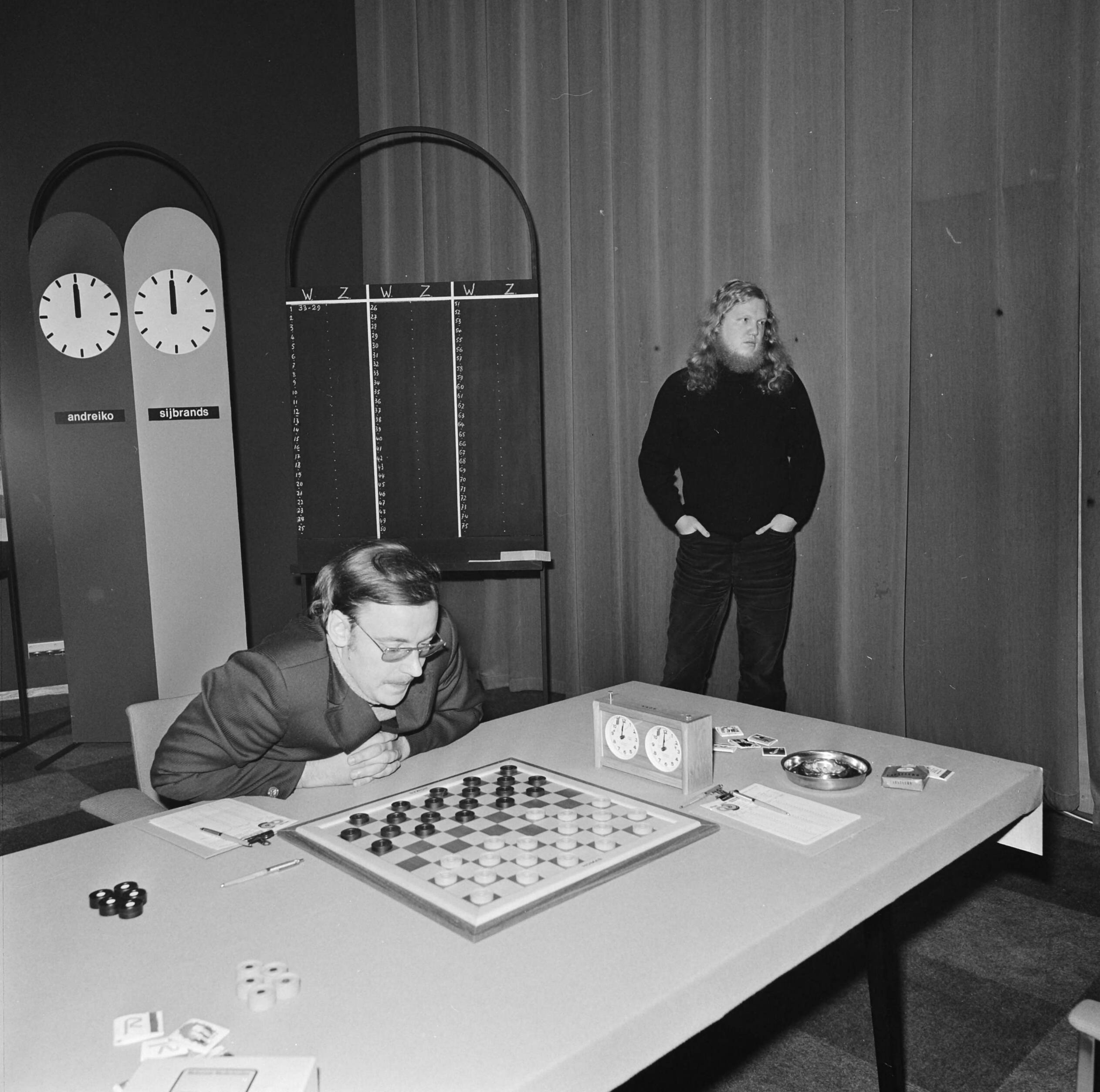 Fotocollectie Anefo — Wereldkampioenschap dammen te Den Haag, Andreiko (gebogen over het bord) en Sijbrands (staand) — Foto: Mieremet, Rob / Anefo. Licentie: Set B: Rechtenvrij / Publiek Domein. Bron: Nationaal Archief. http://hdl.handle.net/10648/ac384dca-d0b4-102d-bcf8-003048976d84