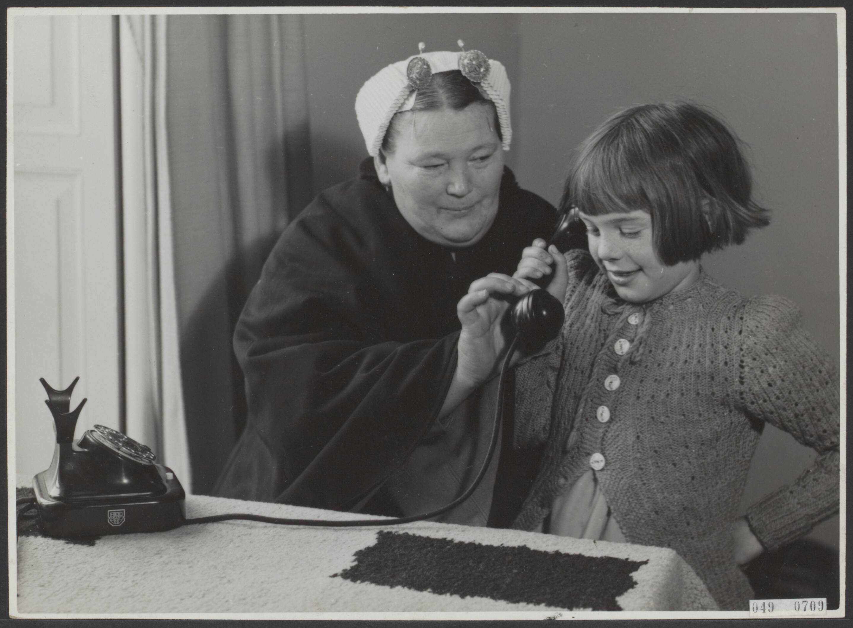 Fotocollectie Elsevier Binnenland — [Jong meisje houdt telefoon vast. Naast haar een vrouw in Scheveningse klederdracht] — Foto: Associated Correspondents. Licentie: Set B: Rechtenvrij / Publiek Domein. Bron: Nationaal Archief. http://hdl.handle.net/10648/ae01fa20-d0b4-102d-bcf8-003048976d84