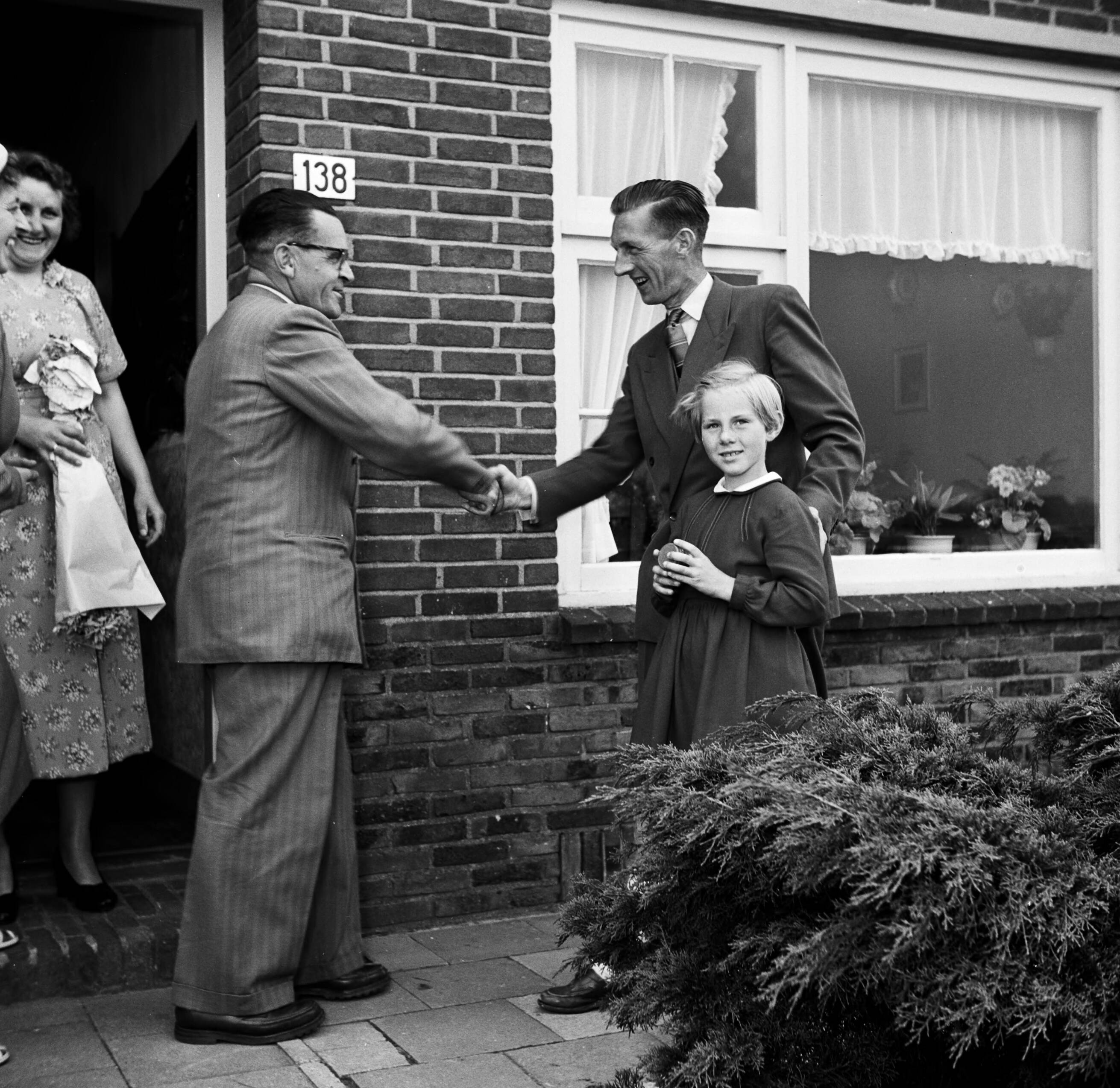 Fotocollectie Van de Poll — Gerard de Jong bij de voordeur van zijn woning, schudt de hand van Ynse Postma. Links zijn echtgenote en naast hem zijn dochterje Marietje — Foto: Poll, Willem van de. Licentie: Set B: Rechtenvrij / Publiek Domein. Bron: Nationaal Archief. http://hdl.handle.net/10648/aecce168-d0b4-102d-bcf8-003048976d84